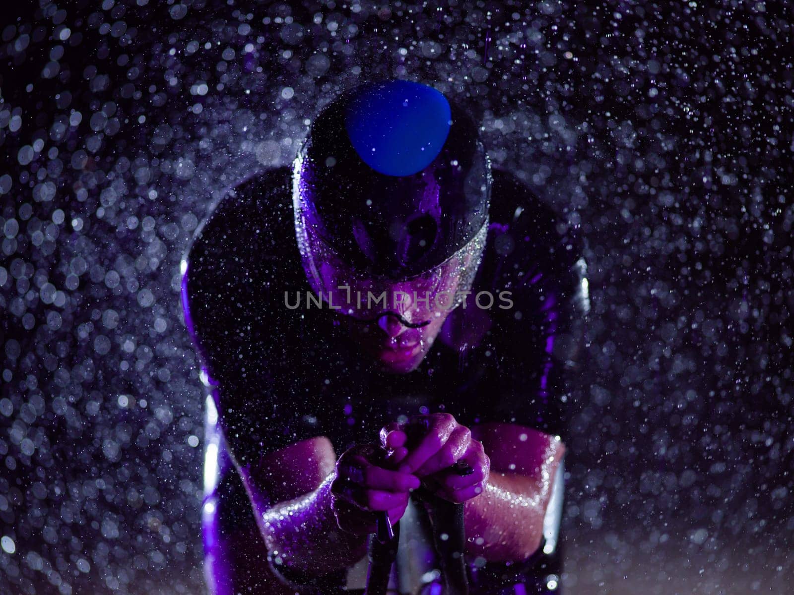 A triathlete braving the rain as he cycles through the night, preparing himself for the upcoming marathon. The blurred raindrops in the foreground and the dark, moody atmosphere in the background add to the sense of determination and grit shown by the athlete