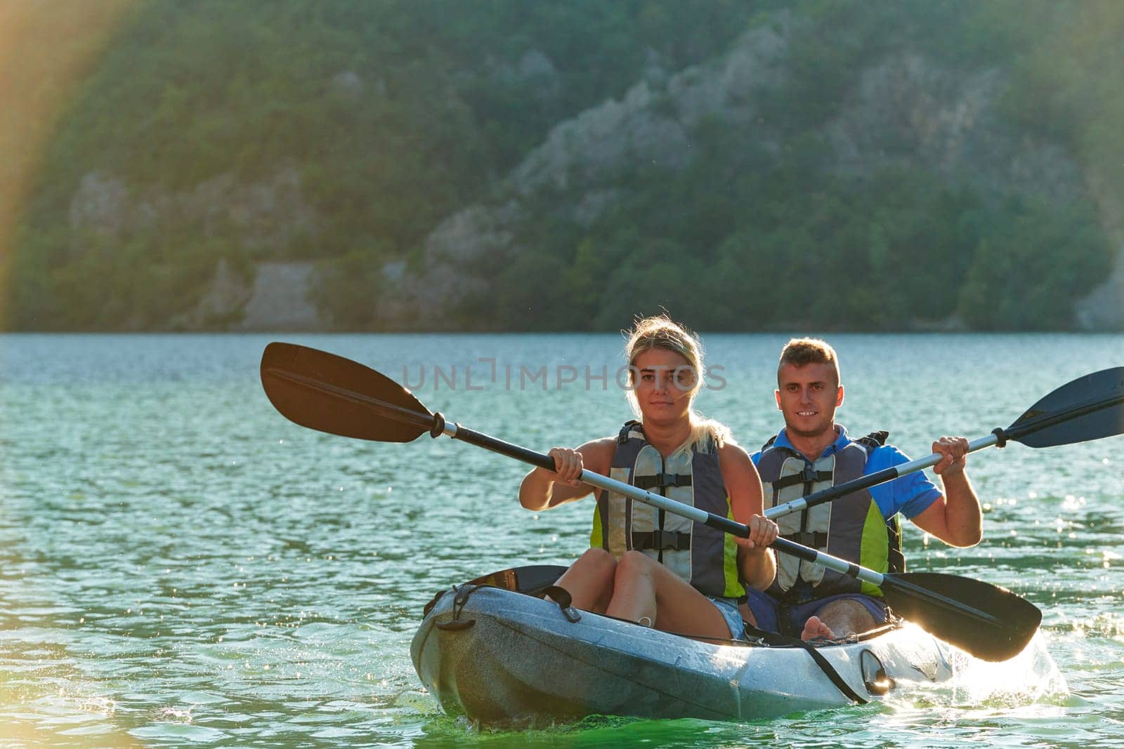 A young couple enjoying an idyllic kayak ride in the middle of a beautiful river surrounded by forest greenery in sunset time.