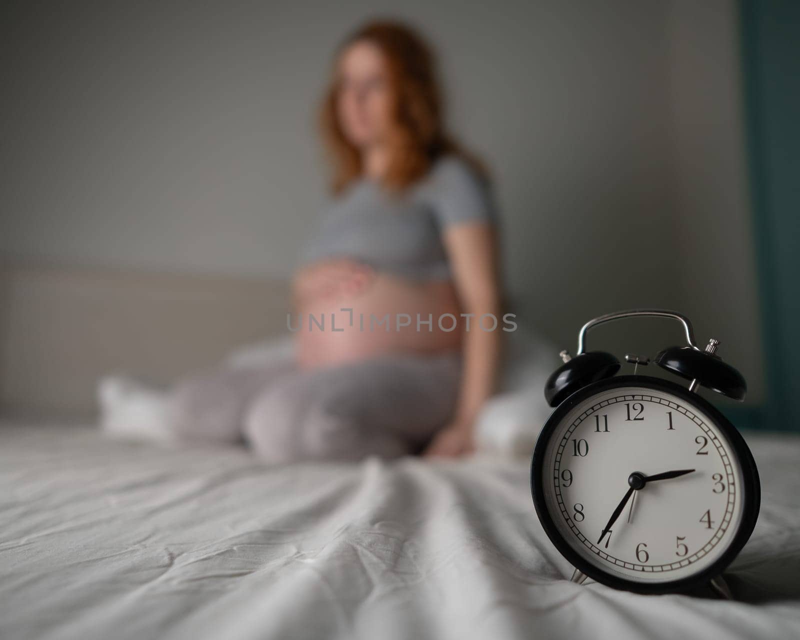 Caucasian pregnant woman sits on the bed and suffers from insomnia. Alarm clock in the foreground. by mrwed54