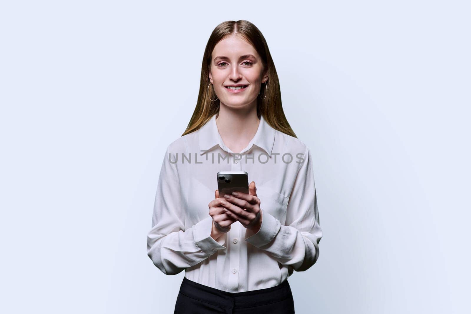 Teenage student girl 16, 17, 18 years old in white shirt holding smartphone in hands, smiling looking at camera on white studio background. Education, technology, high school, college, lifestyle, youth concept