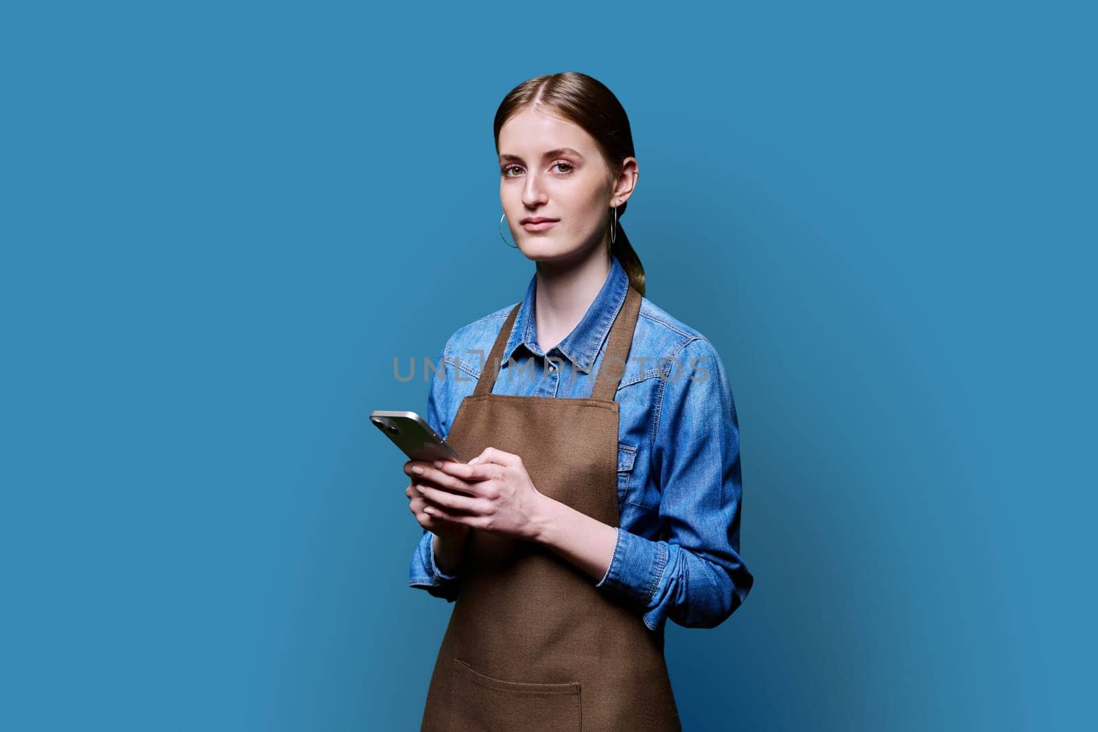 Young female worker in an apron looking at camera holding smartphone on blue studio background. Work, business, online internet services, mobile apps applications, technology concept