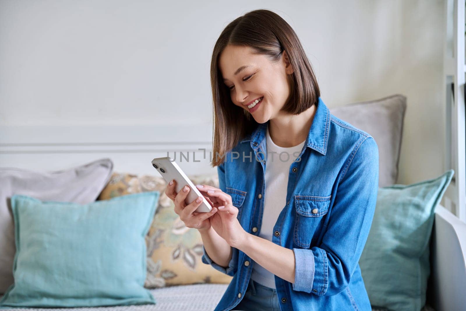 Young woman using smartphone sitting on couch at home. Female texting reading looking, mobile applications technology internet online services for work study leisure communication shopping lifestyle