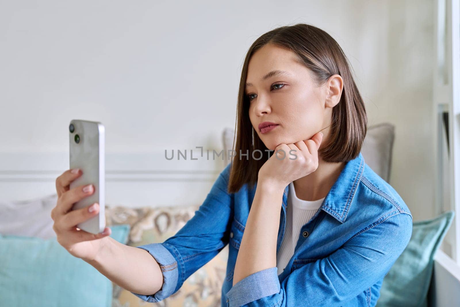 Young serious woman sitting with smartphone on couch at home having video call. Female chatting with friends colleagues on social networks, student talking in online lesson, freelancer working remotely