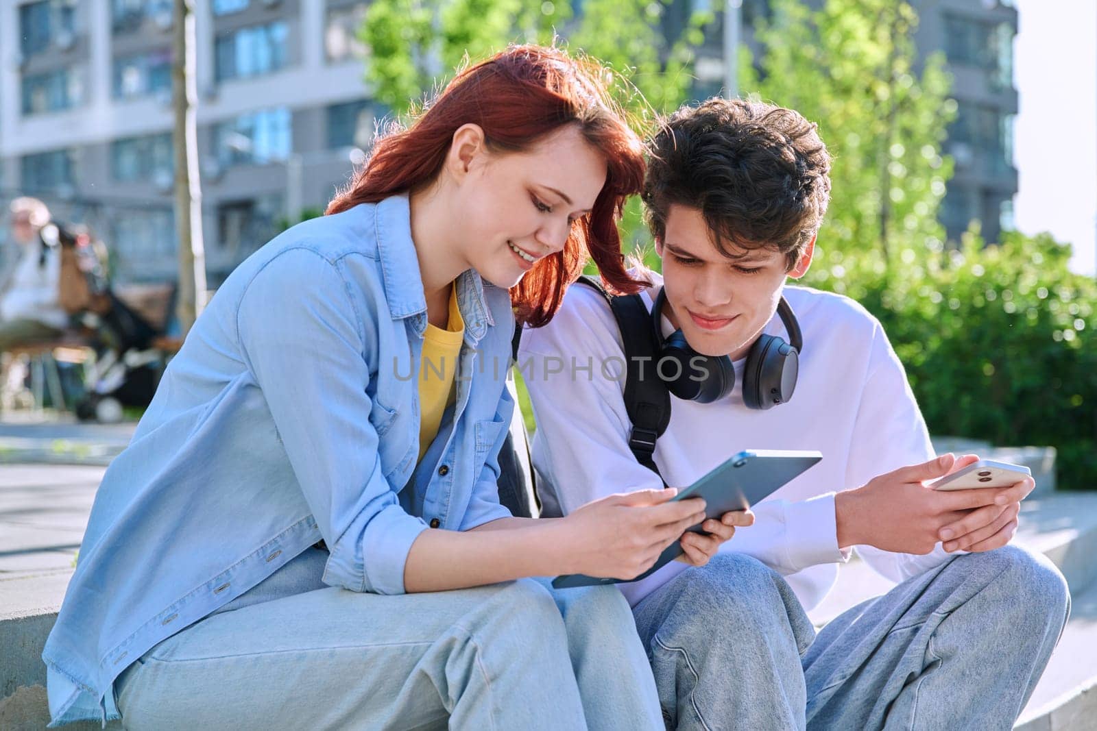 Teenage youth friends guy and girl university college students sitting outdoor on campus steps talking laughing using smartphone. Technology, lifestyle concept