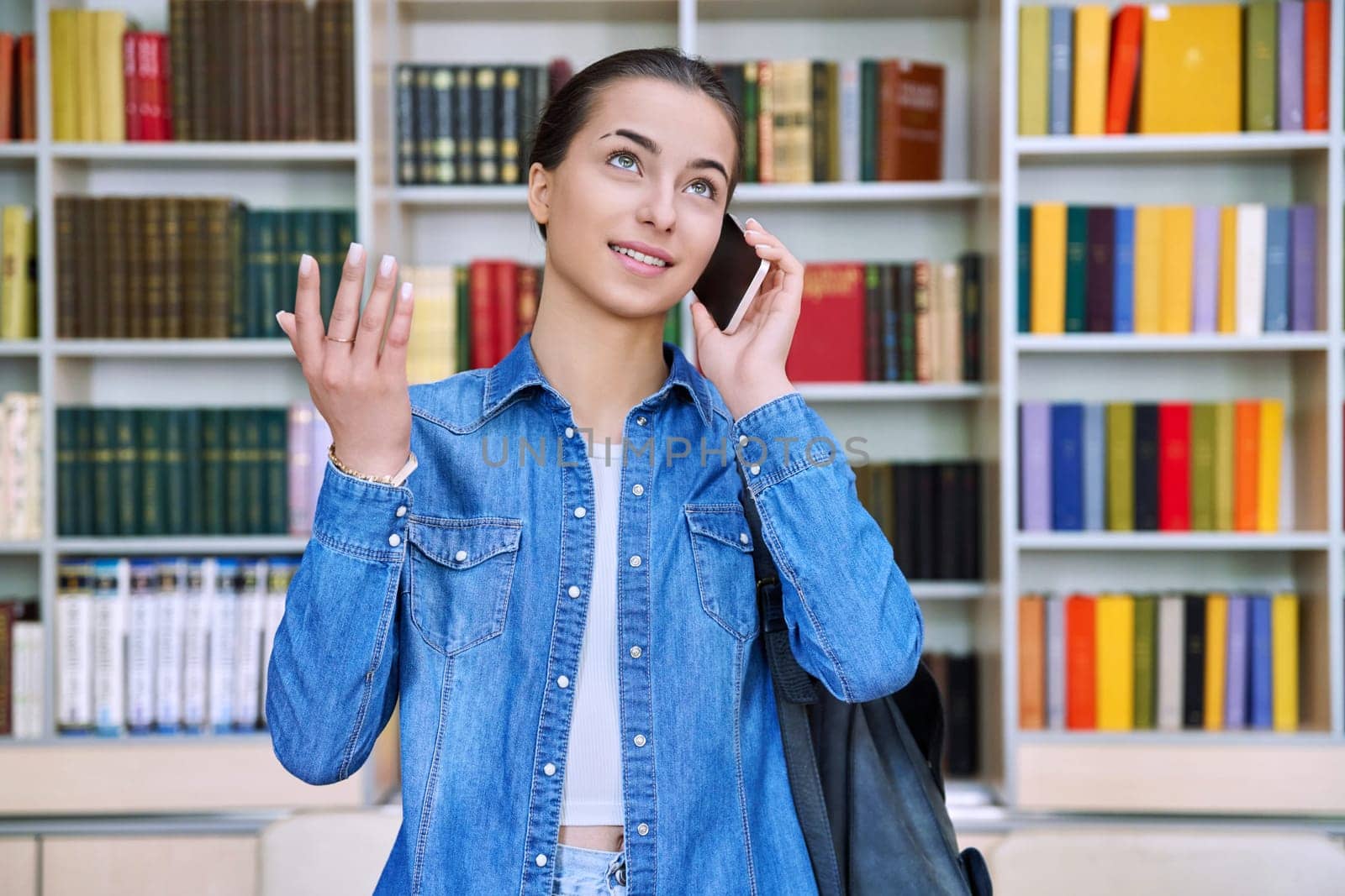 Teenage girl student with backpack talking on mobile phone, inside high school building, in library. Adolescence, education, lifestyle concept