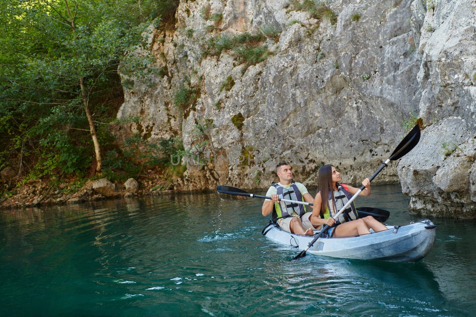 A young couple enjoying an idyllic kayak ride in the middle of a beautiful river surrounded by forest greenery by dotshock