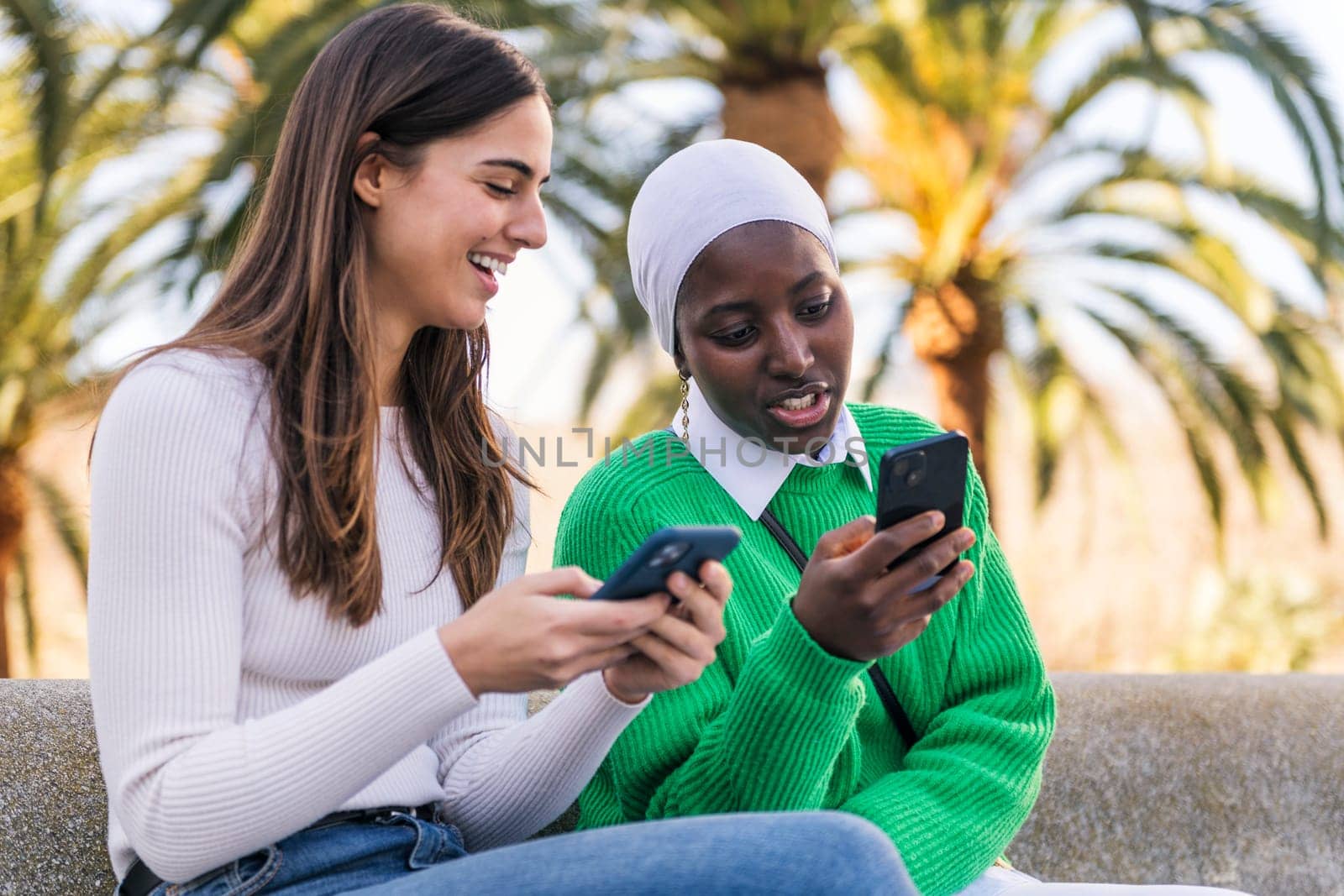 two young female friends using mobile phone sitting in a city park, concept of technology of communication and urban lifestyle