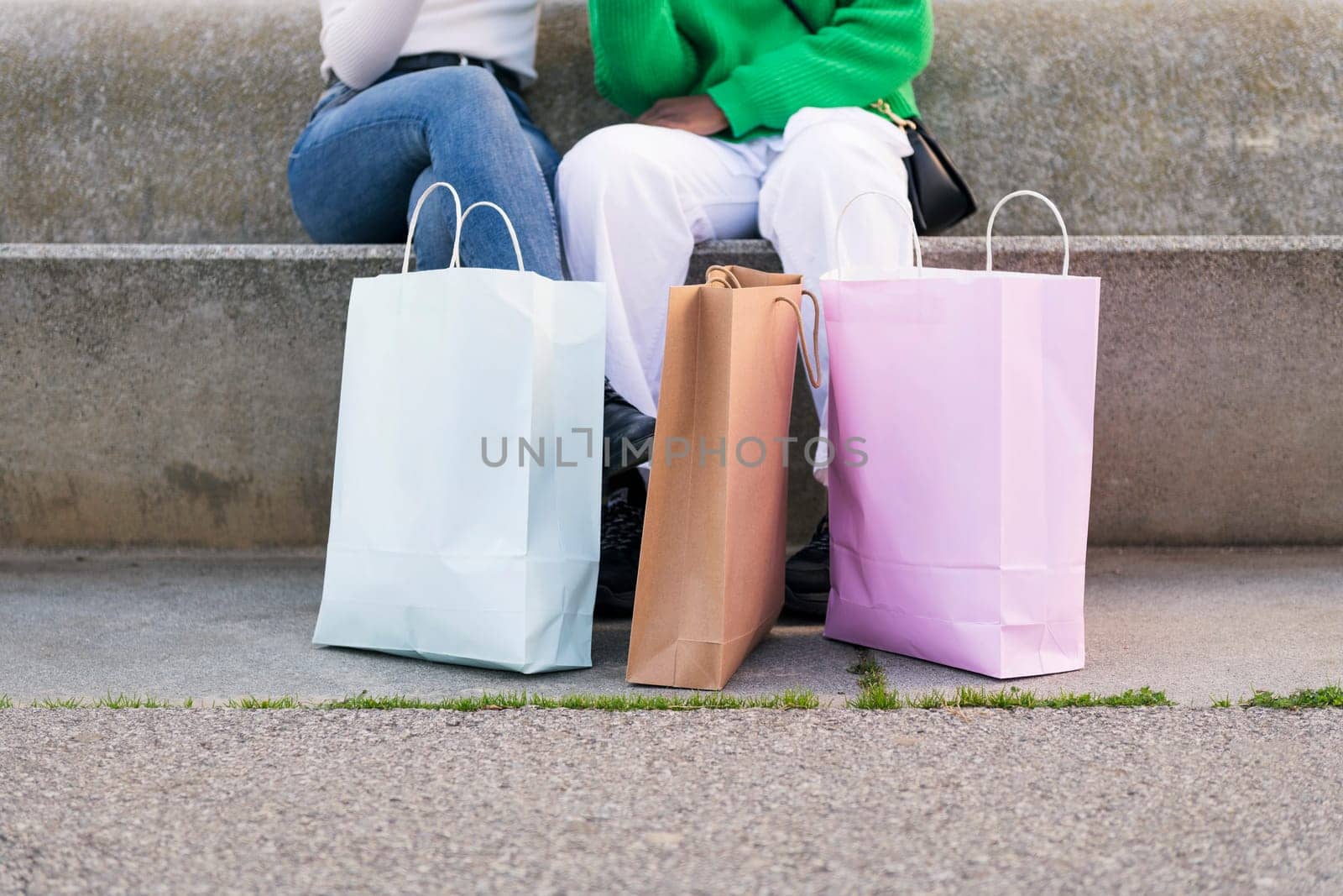 two unrecognizable female friends sitting in a city park with shopping bags, concept of buying and urban lifestyle