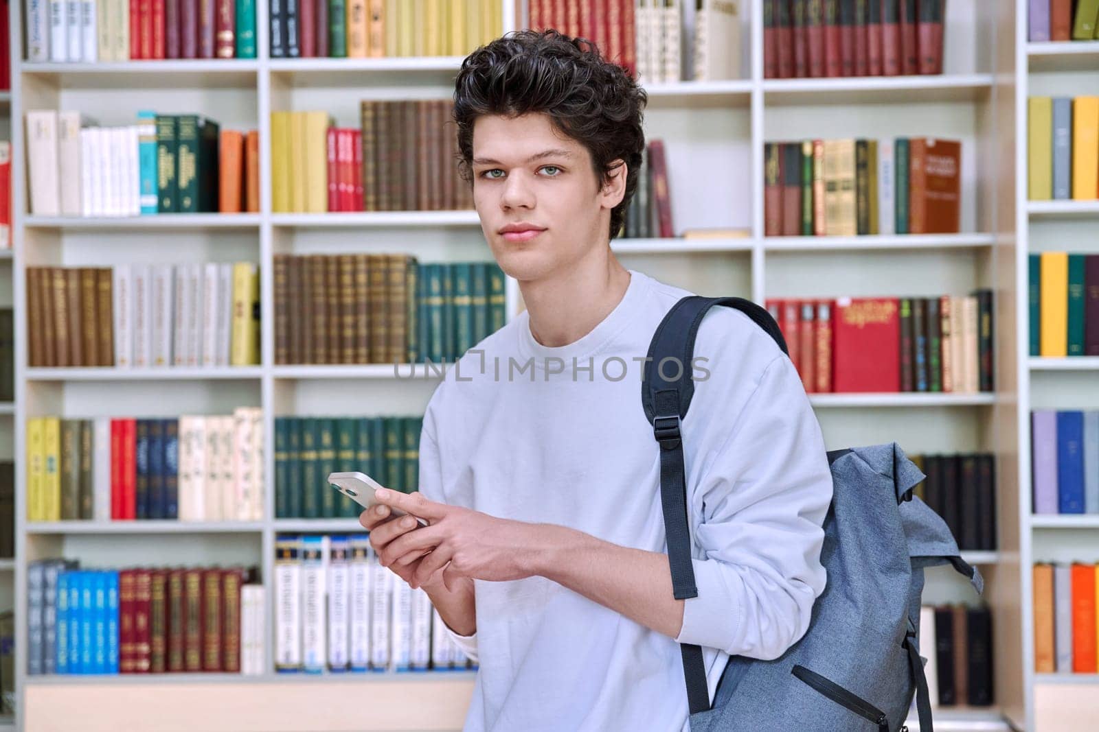 Teenage male student with backpack using smartphone, inside college building, in library. Technologies, mobile educational apps applications, services, e-learning concept