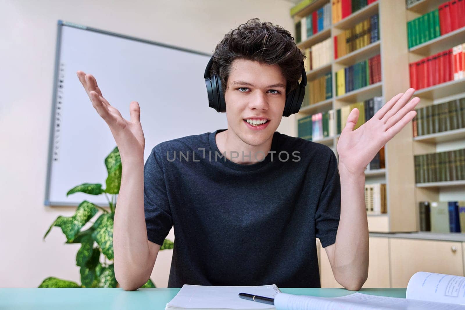 Web cam portrait of college student guy in headphones looking talking to camera in educational building library classroom. Video call chat conference, online lesson exam test, technology education