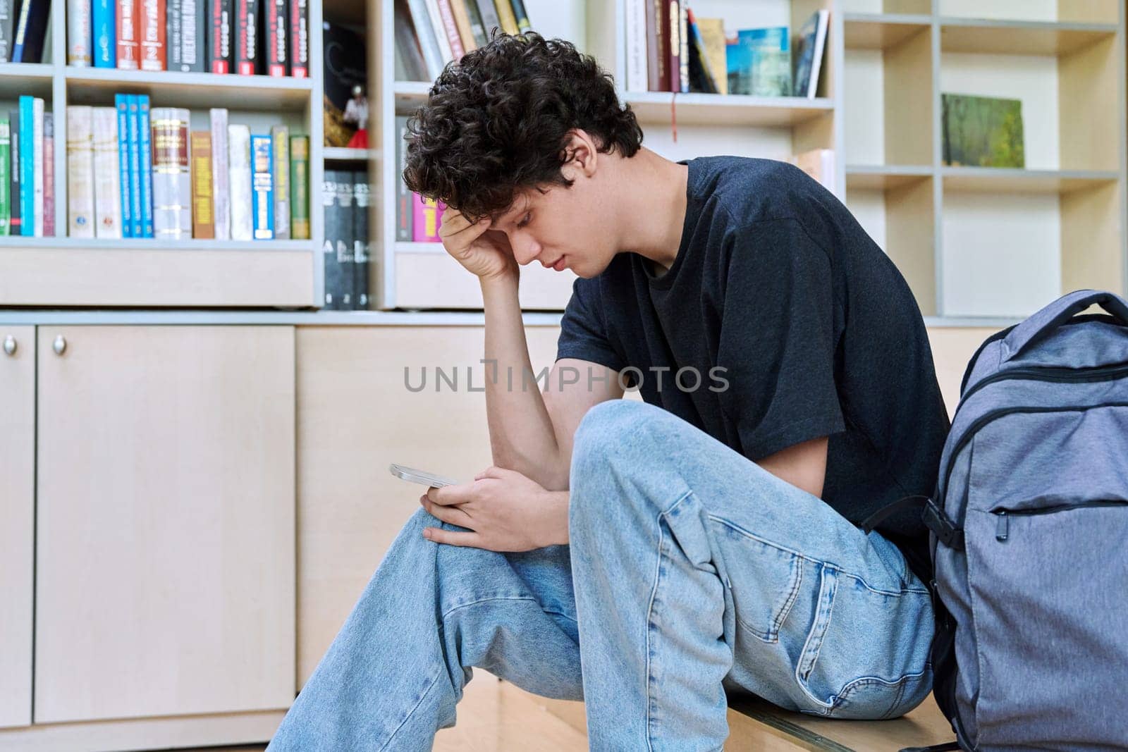 Sad upset young male college student looking at smartphone while sitting in classroom. Negative emotions, difficulties, troubles, youth concept