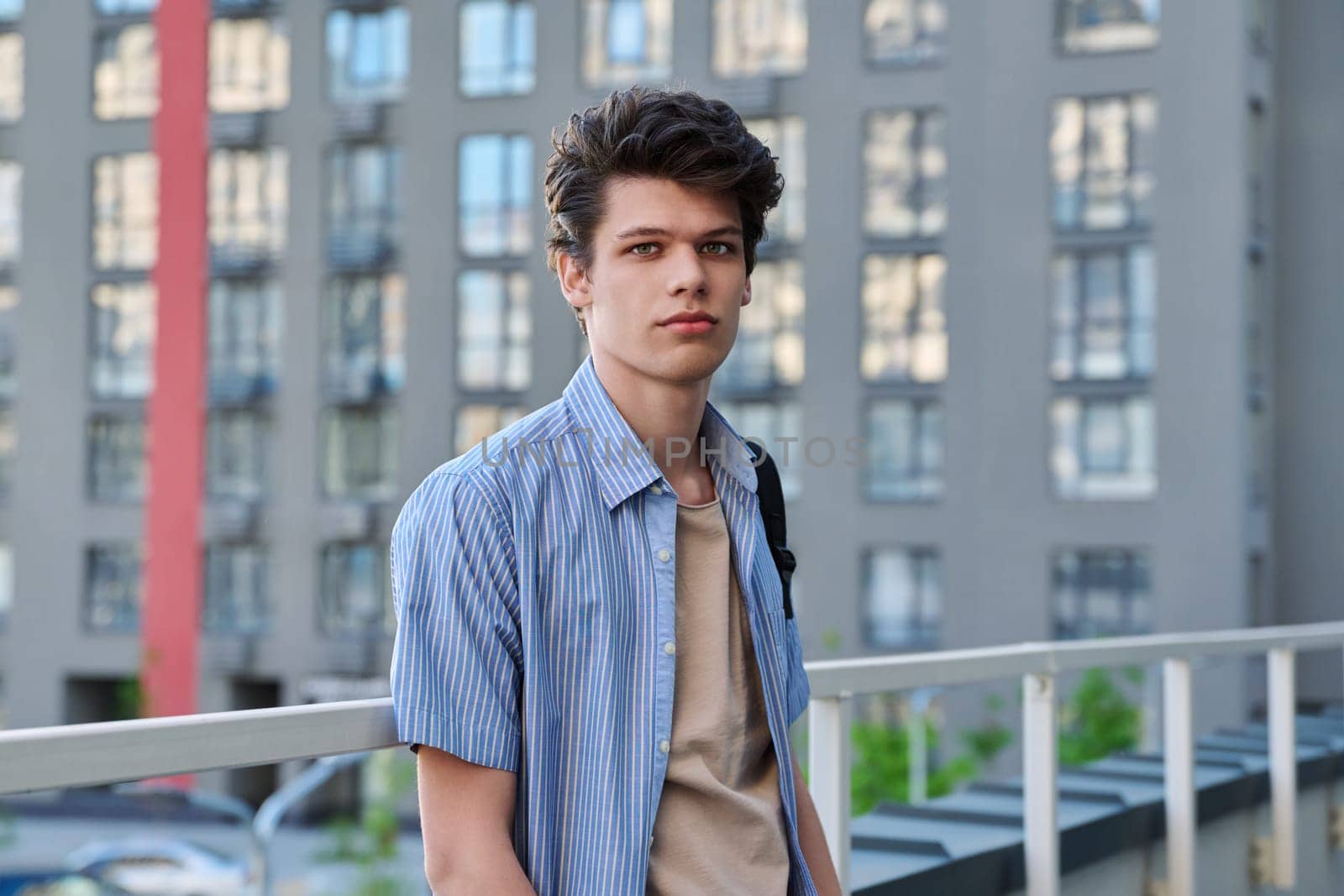 Portrait of smiling, handsome student guy 19, 20 years old, looking at camera outdoors, modern building background. Youth, urban style, young man, lifestyle concept