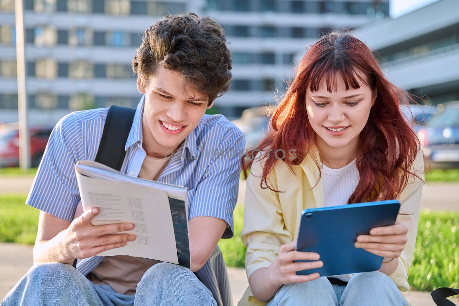 Teenage college students guy and girl talking, using digital tablet, sitting outdoor, urban modern city. Youth 19-20 years old, education, technologies, lifestyle, friendship concept