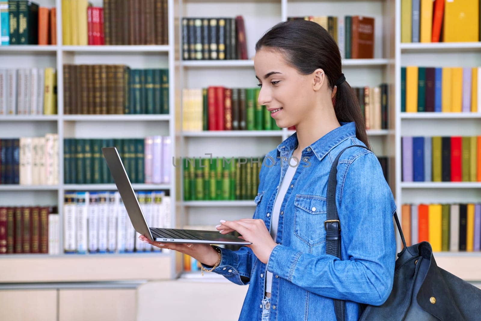 Teenage girl student with backpack using laptop computer, inside high school building, in library. Technologies, internet mobile educational apps applications, services, e-learning concept