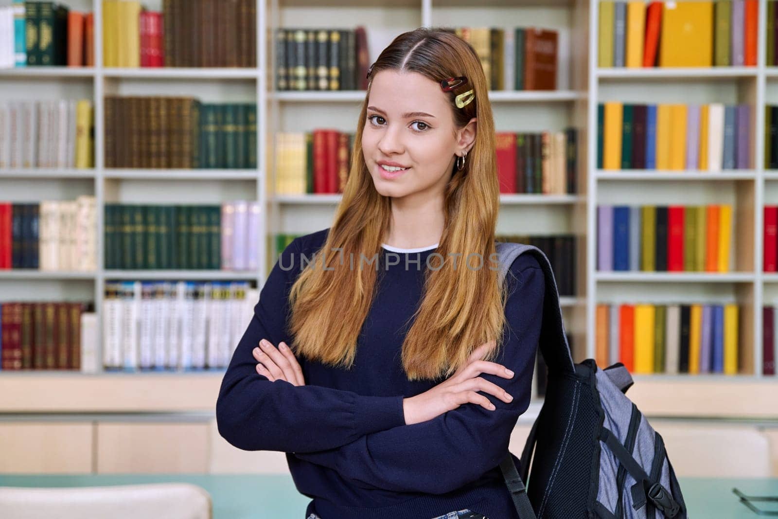 Portrait of high school student girl with backpack in library classroom. Red-haired teenage female 16, 17 years old looking at camera. Education, adolescence, school concept