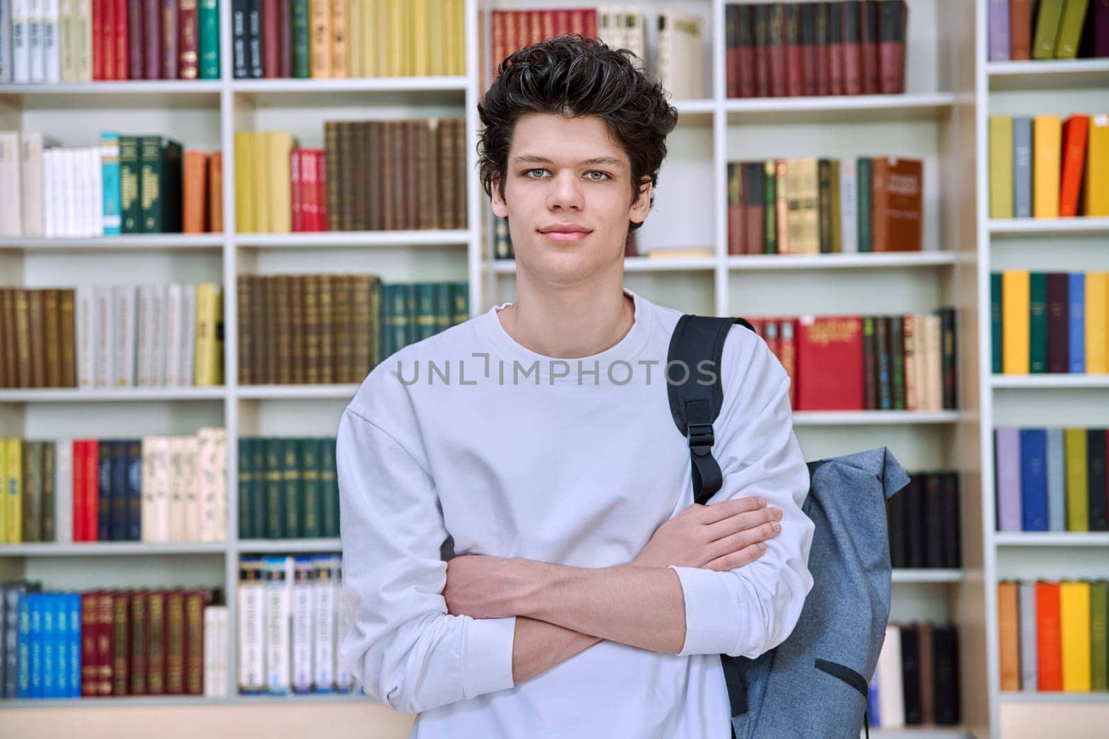 Portrait of confident handsome college student guy with crossed arms inside library of educational building. Education, youth, lifestyle concept