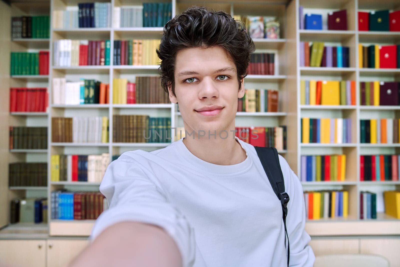 Selfie portrait of smiling handsome college student guy inside library of educational building. Education, youth, lifestyle concept