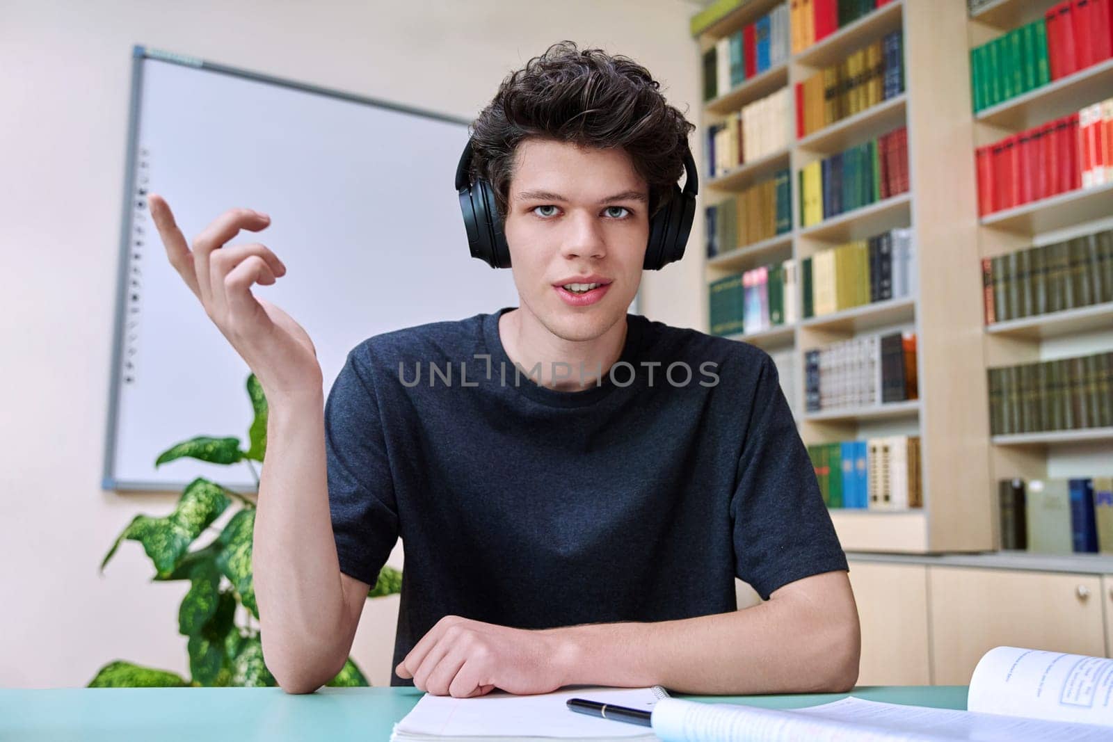 Web cam portrait of college student guy in headphones looking talking to camera in educational building library classroom. Video call chat conference, online lesson exam test, technology education