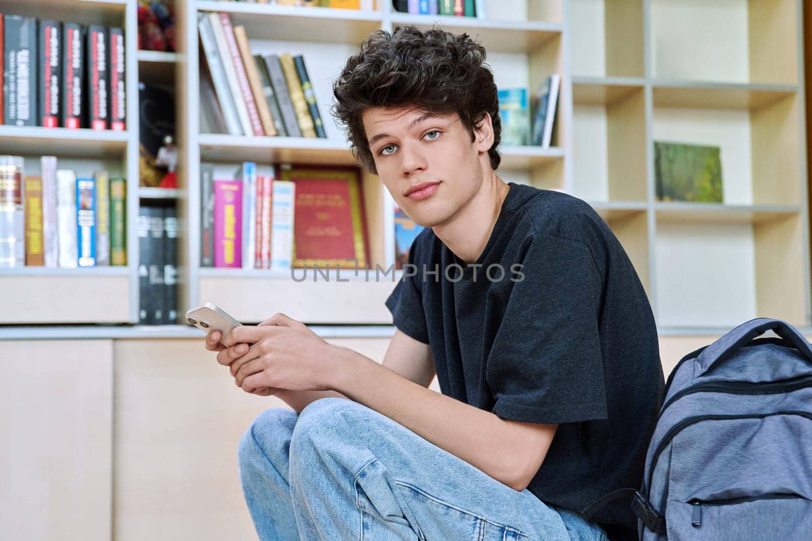 Relaxed handsome young male college student using smartphone, sitting inside library classroom. Teenager guy 19-20 years old looking at camera, education technology mobile applications lifestyle youth