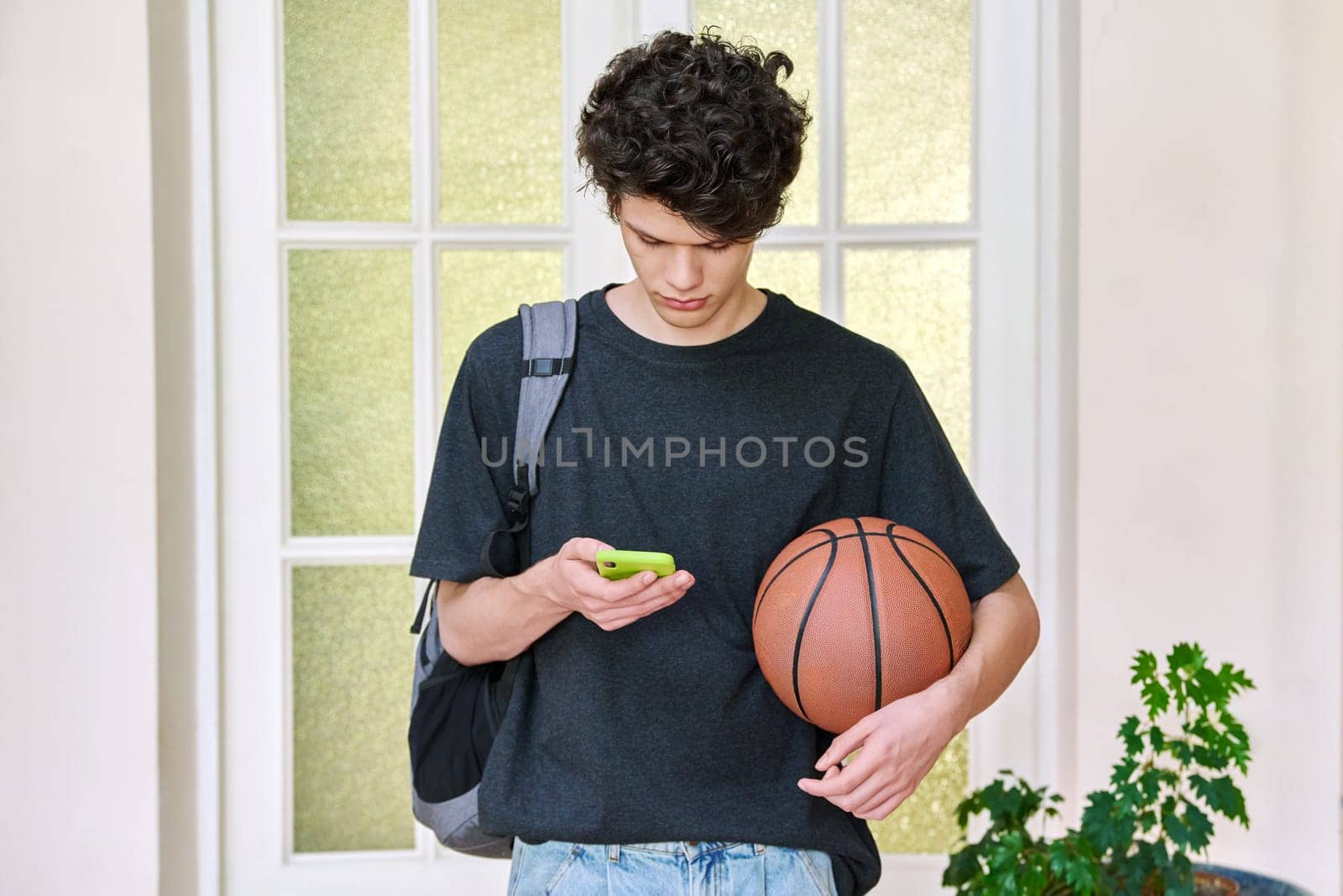 Portrait of young male with backpack, basketball and smartphone. Handsome curly 19, 20 year old teen student using texting mobile phone inside college. Lifestyle, youth concept