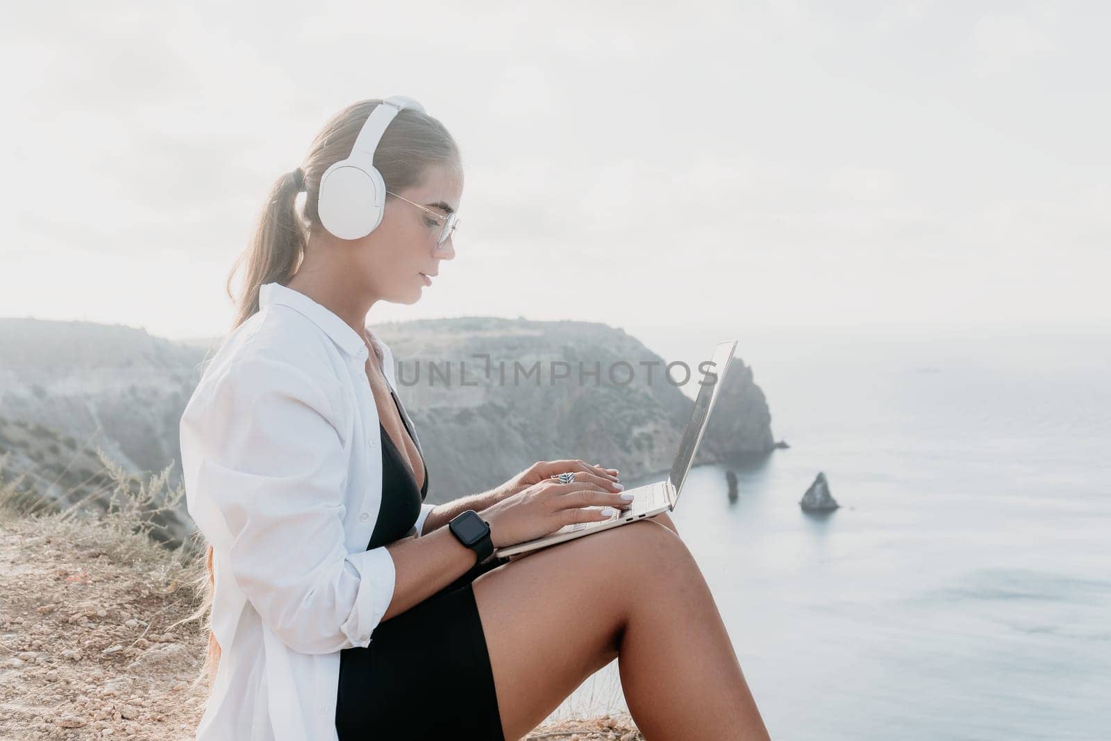 Digital nomad, woman in the hat, a business woman with a laptop sits on the rocks by the sea during sunset, makes a business transaction online from a distance. Freelance, remote work on vacation.