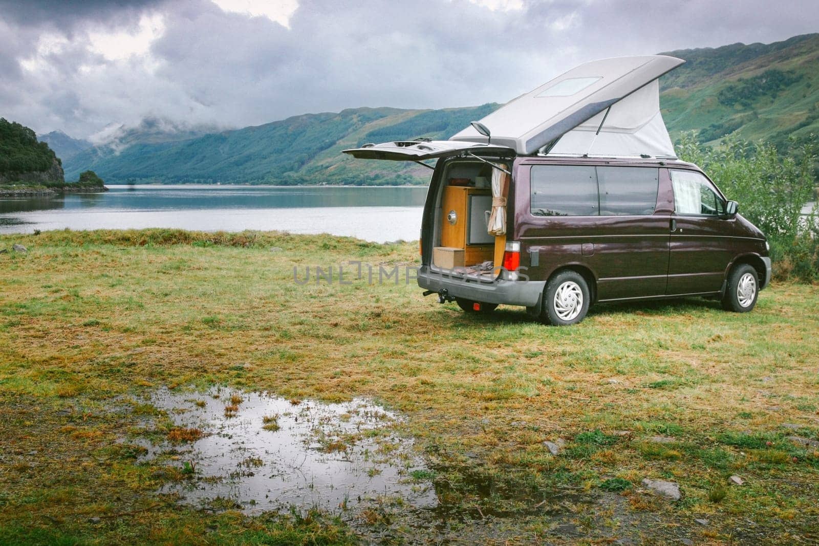 A brown campervan is parked in a grassy field next to a lake. The van has an open roof and the sky is cloudy. The scene has a calm and serene atmosphere, with the lake. Road trip concept.