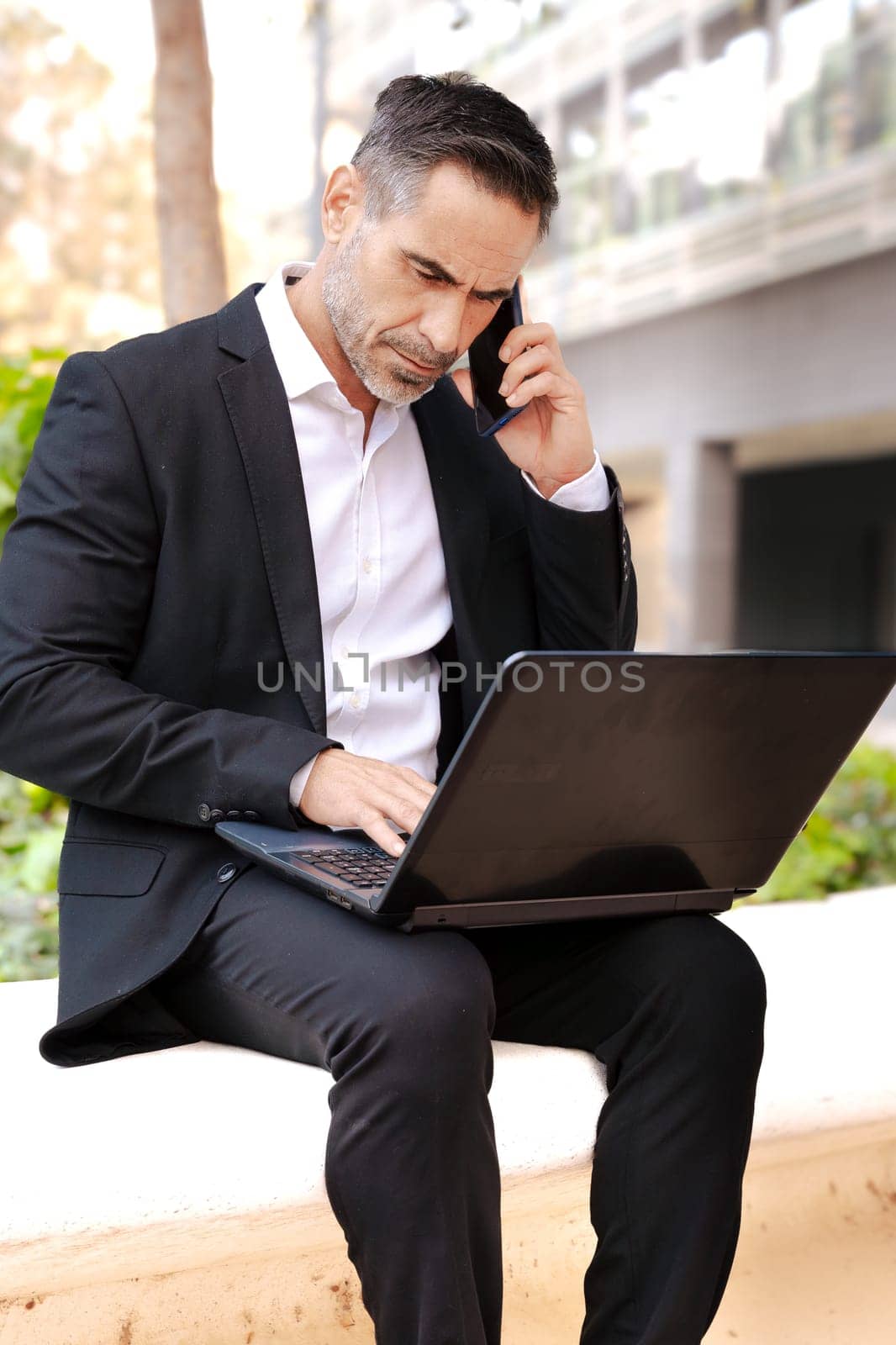 Businessman talks on a smartphone while working with laptop outdoors. by mariaphoto3