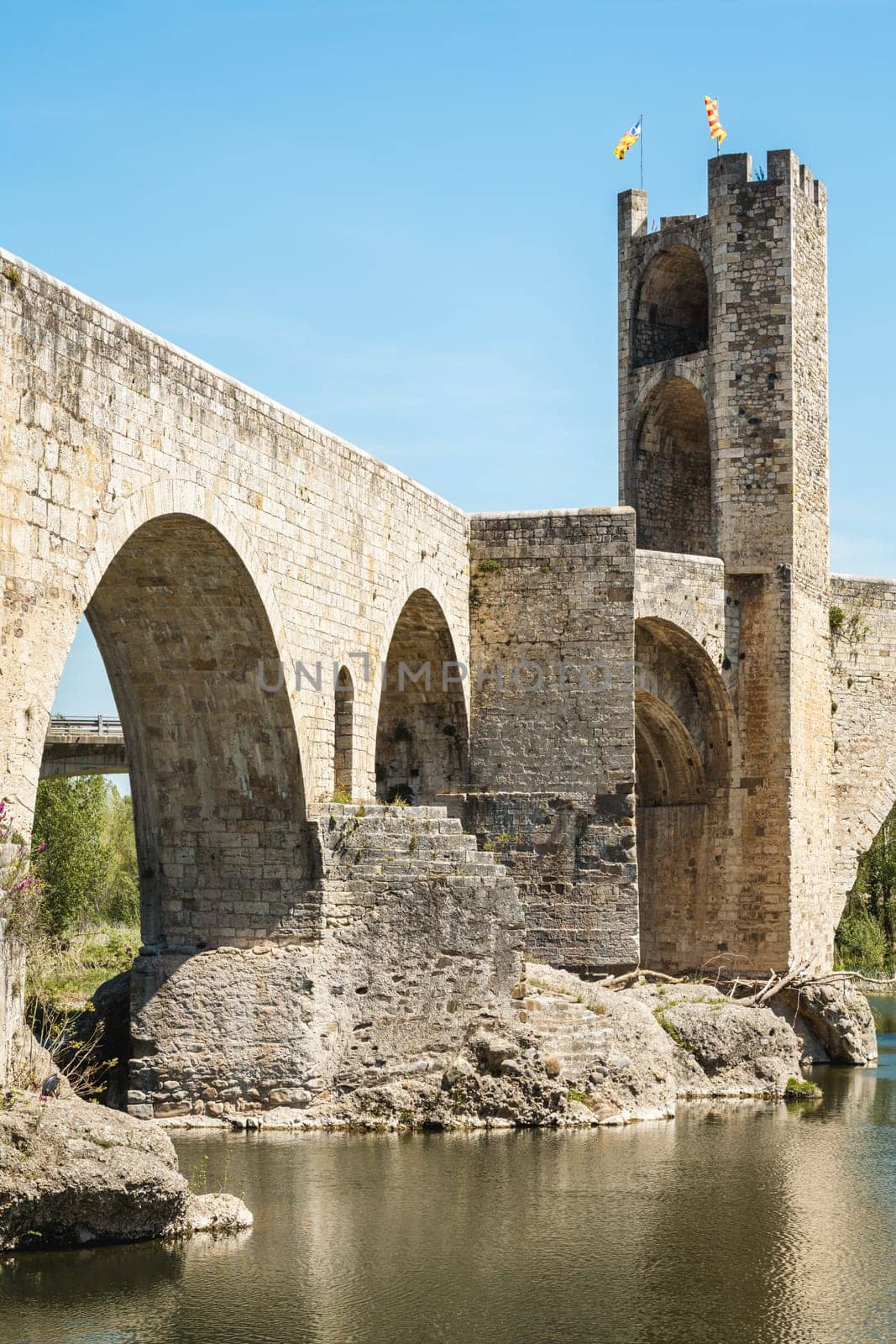 The bridge of the medieval village of Besalú with a tower in the background. The bridge is made of stone and has a stone arch. The water is calm and the sky is clear.