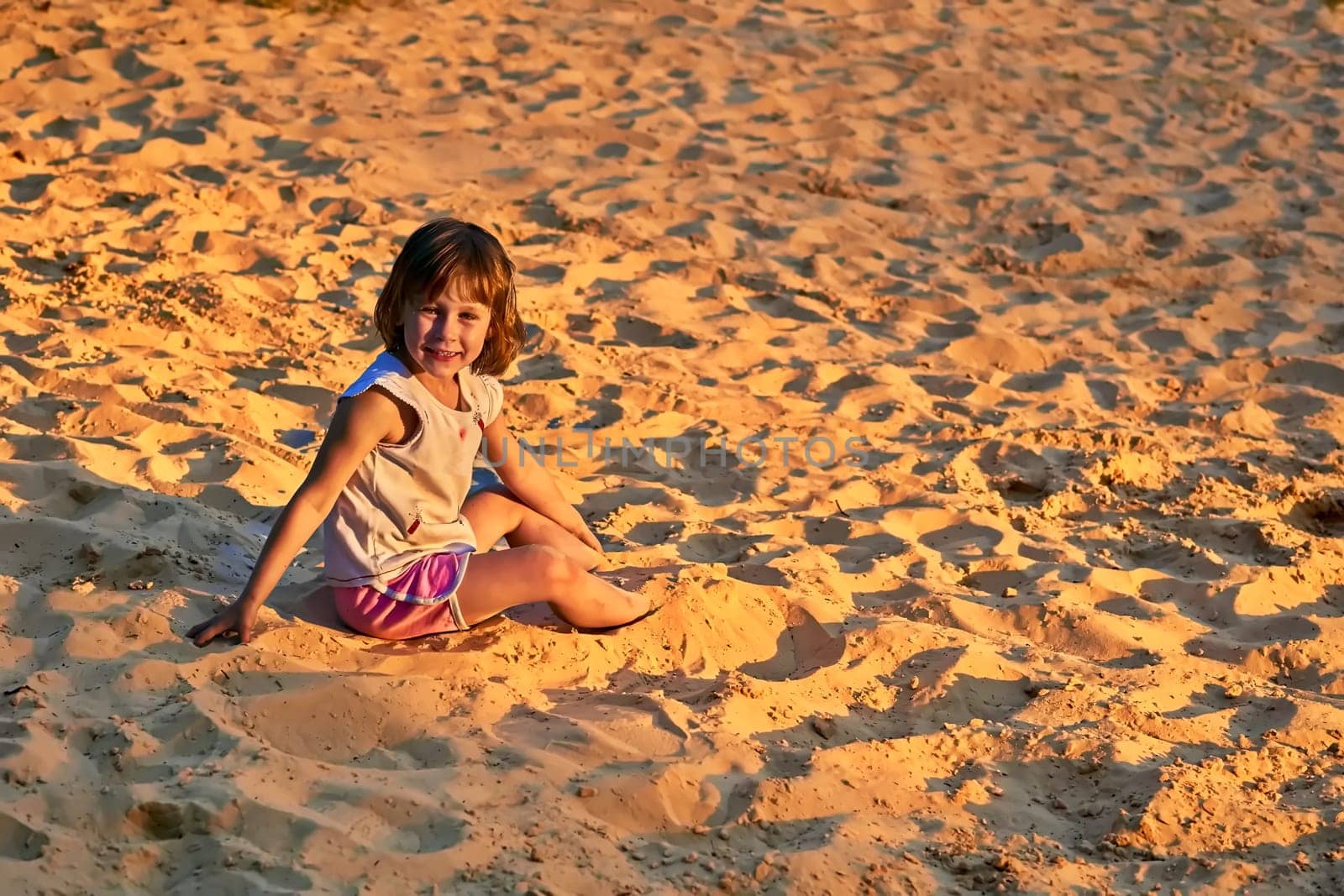 A satisfied smiling girl in a t shirt and shorts sits on beach sand. sunset by jovani68
