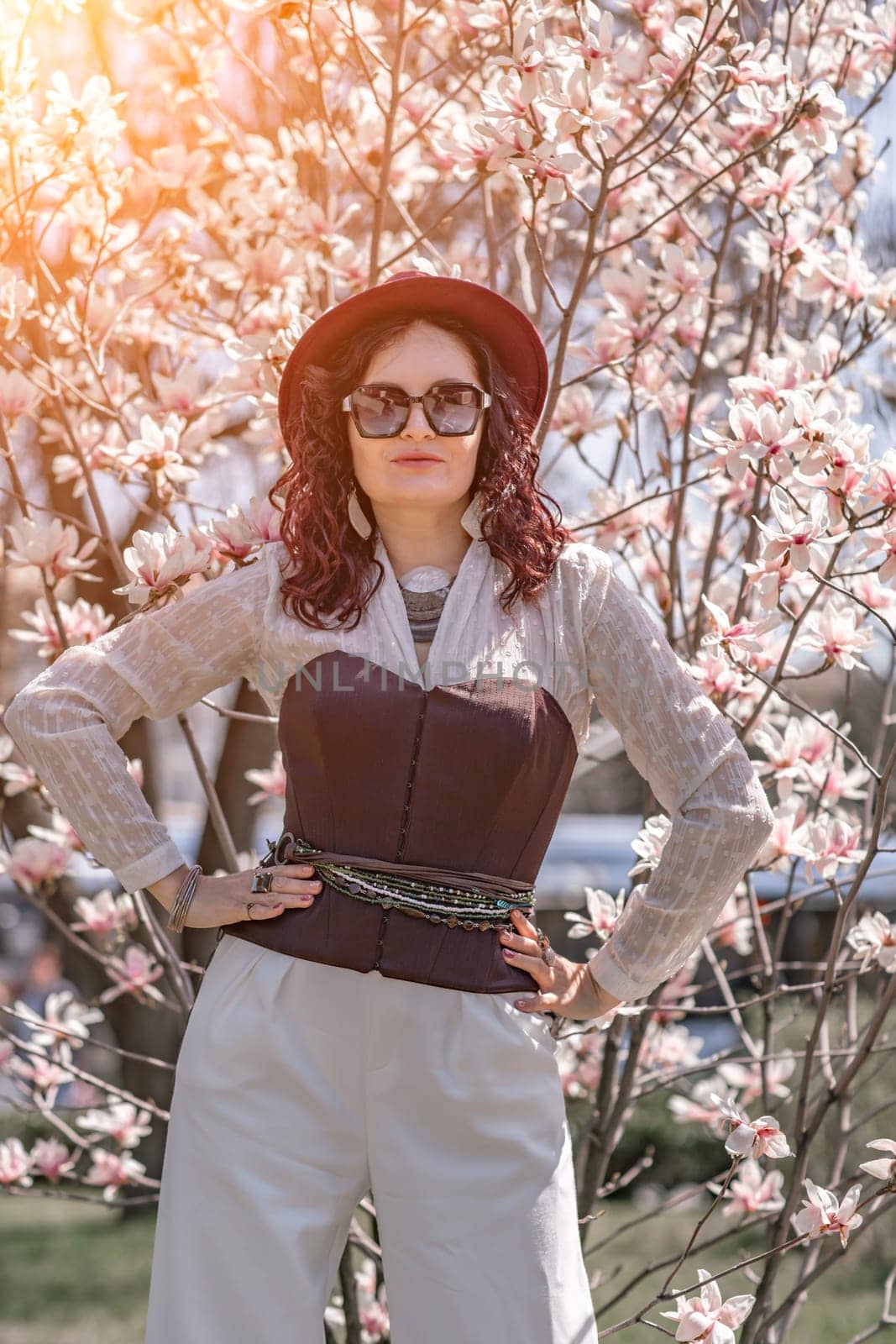 Magnolia park woman. Stylish woman in a hat stands near the magnolia bush in the park. Dressed in white corset pants and posing for the camera. by Matiunina