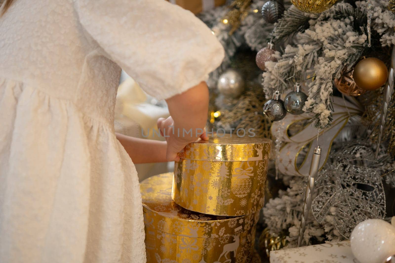 A woman is opening a gold box with a silver ball inside. The box is decorated with gold and silver ornaments, and the woman is wearing a white dress. The scene is set in a room with a Christmas tree