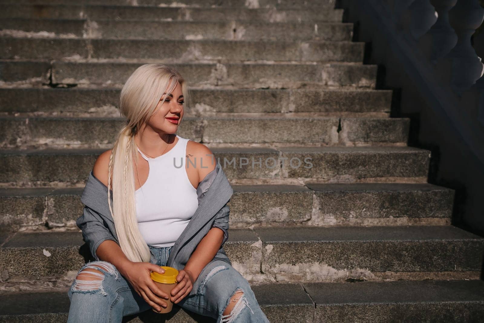 A blonde woman sits on a set of stairs, holding a cup. She is wearing a gray jacket and blue jeans. by Matiunina