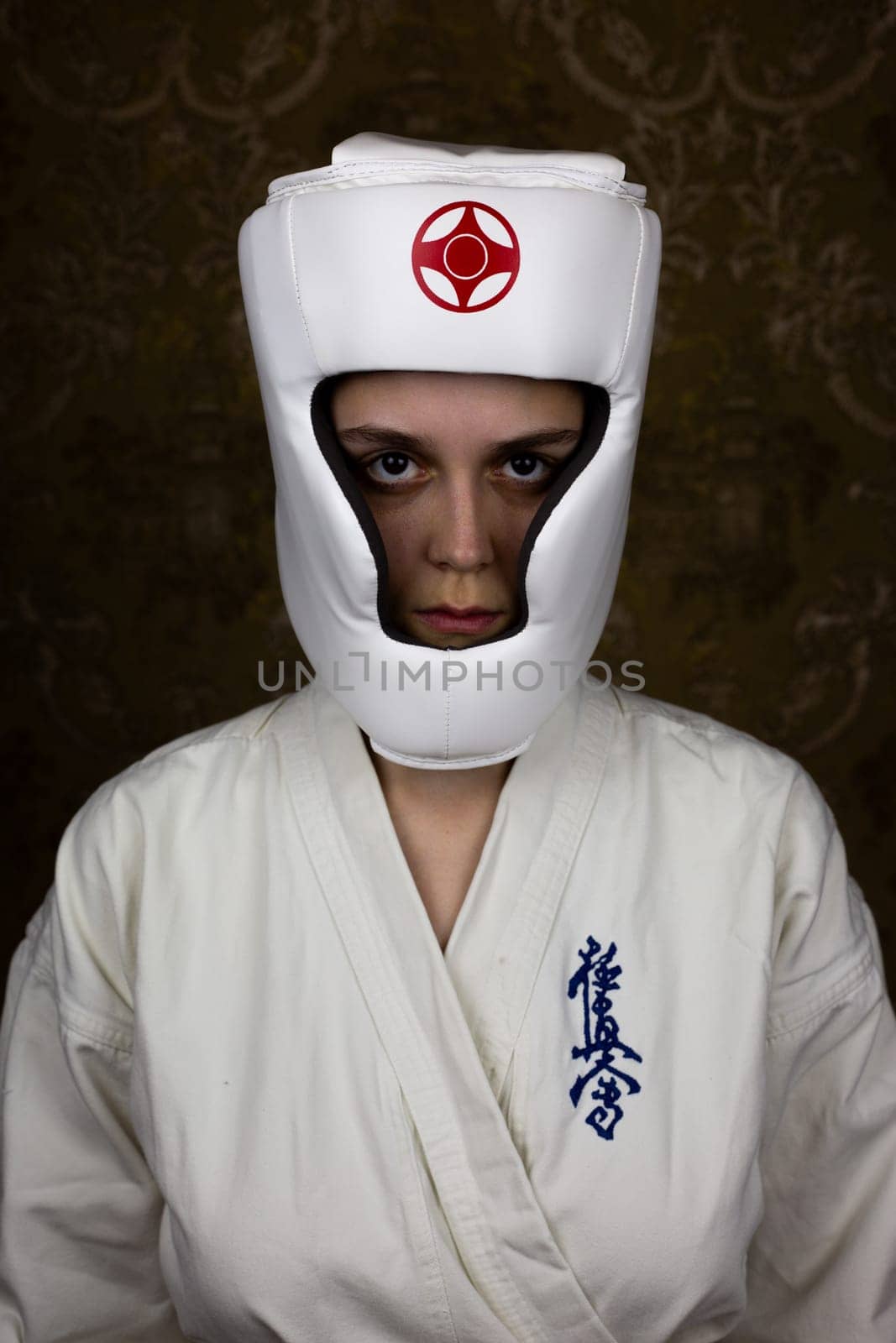 Kyokushinkai karateka girl in protective helmet and kimono looks at the camera, vertical photo of girl in equipment martial arts. Inscription in Japanese: Kyokushinkai