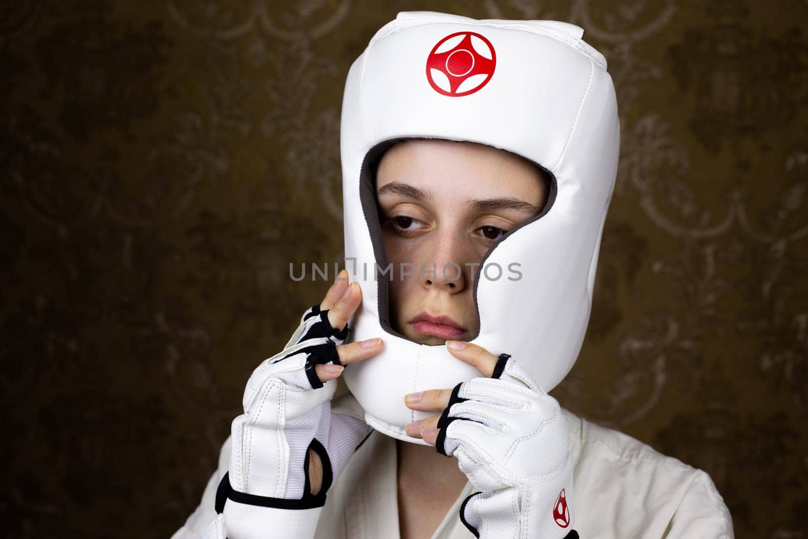 Girl in kimono and special Kyokushin karate gloves puts on protective helmet before fight, safety of martial arts athletes