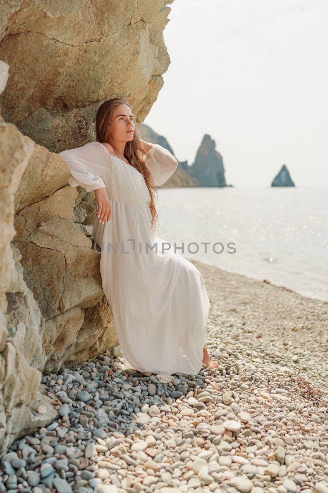 woman white dress standing on a beach, looking out at the ocean. She is lost in thought, with her hand on her hip. Concept of calm and introspection by Matiunina