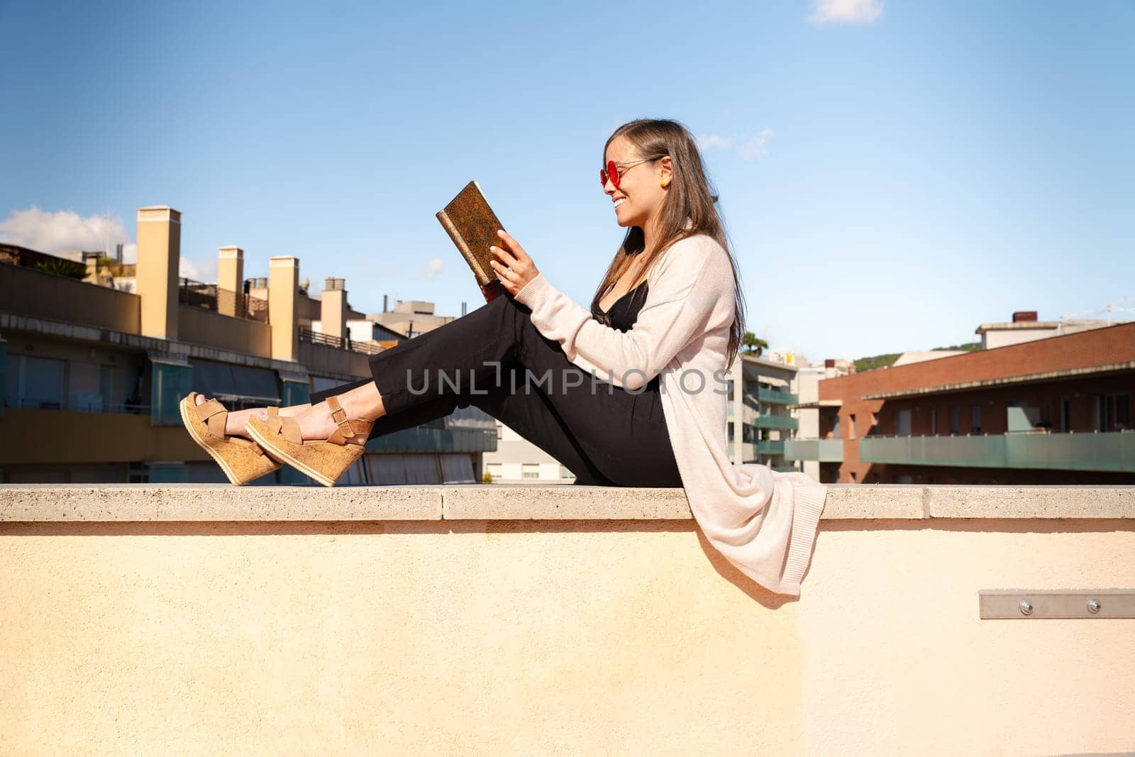 Happy young woman sitting on the roof reading a book outdoors by mariaphoto3