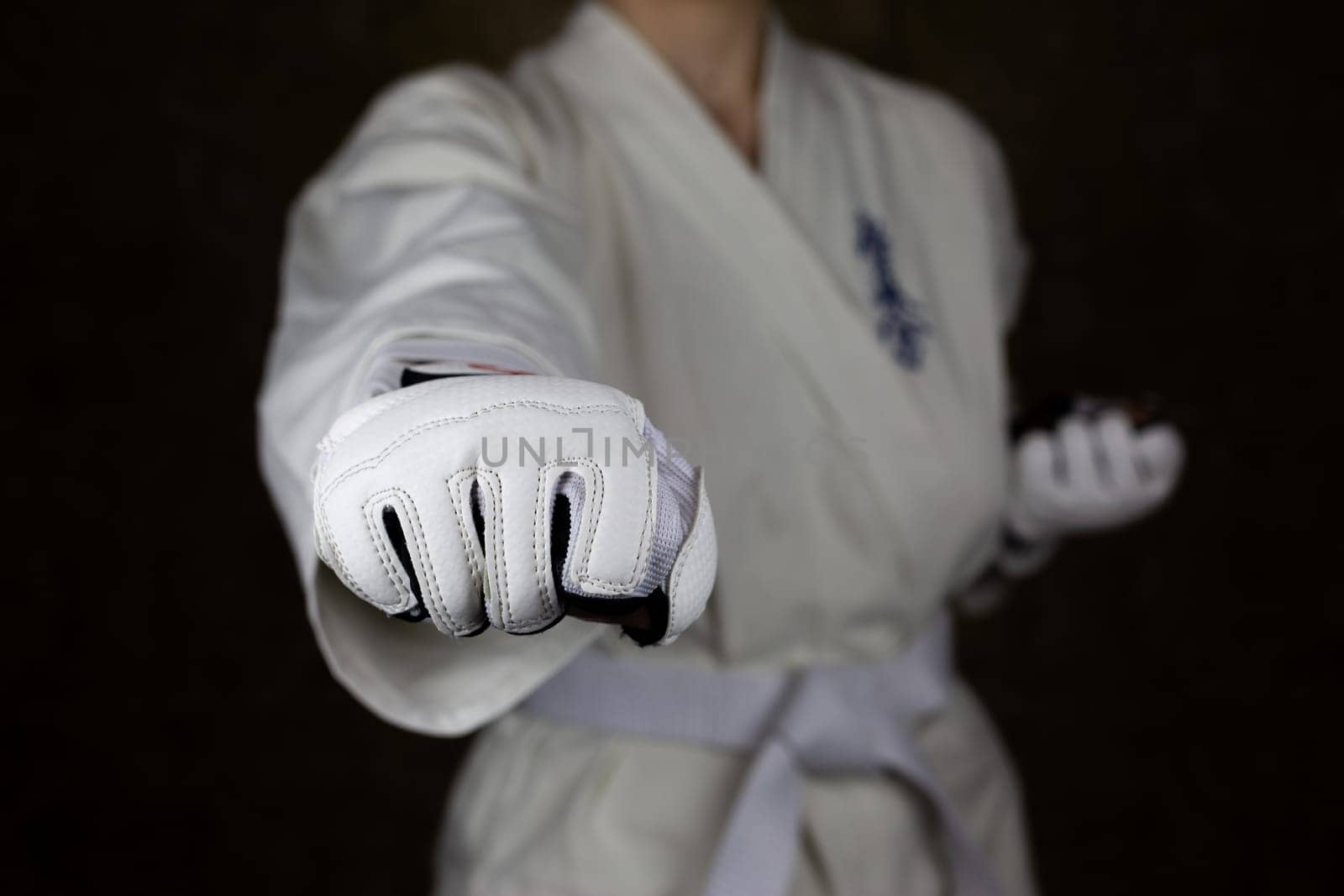 Young woman in kimono and white belt throws punch in protective gloves in Kyokushin karate to camera, focus on fist with blurred martial arts athlete on background. Inscription in Japanese: Kyokushinkai