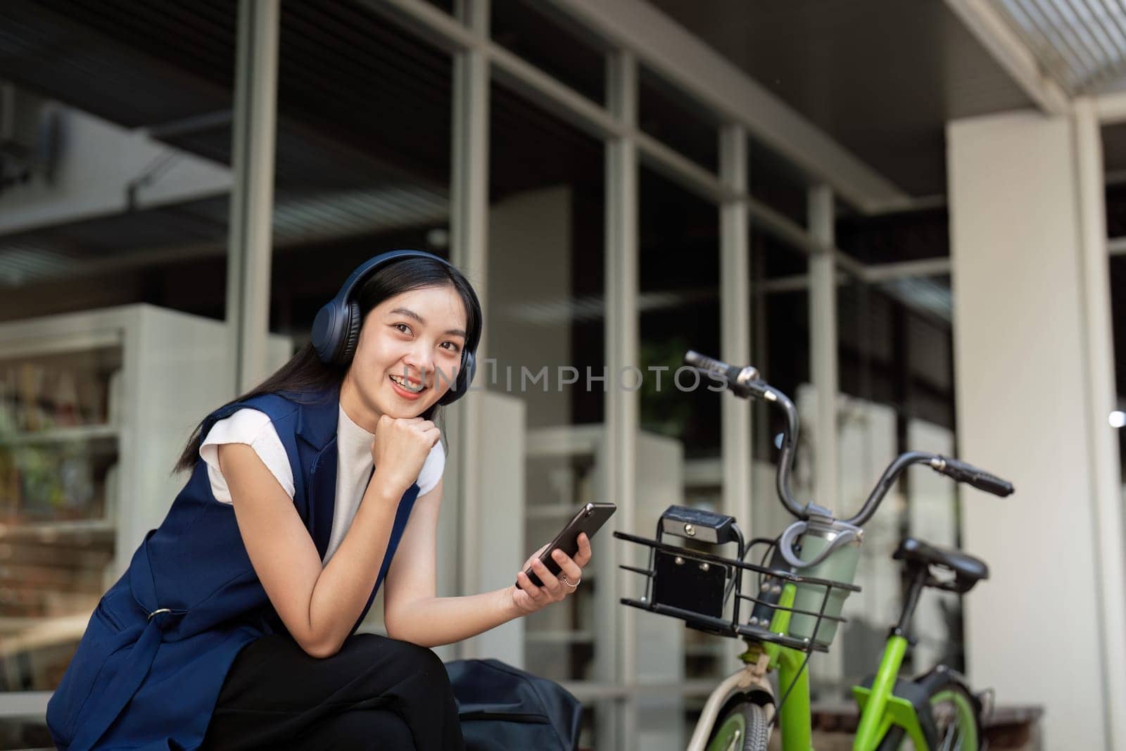 Smiling businesswoman with headphones resting with her bicycle outside office. Concept of relaxation, urban commuting, and modern lifestyle.