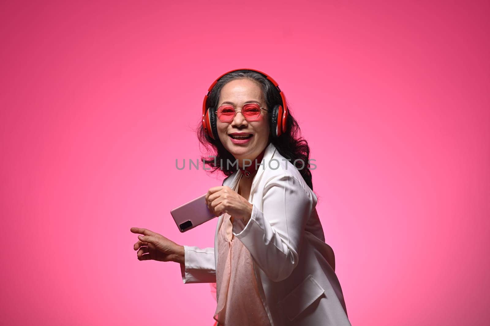 Happy senior woman woman listening to music and dancing on light pink background.