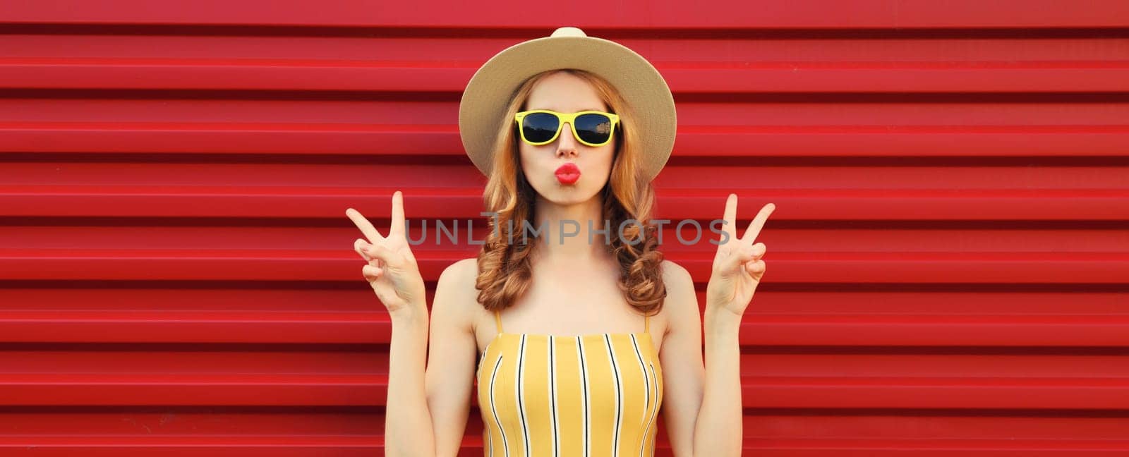 Portrait of beautiful stylish young woman blowing kiss in summer hat, yellow sunglasses posing on red wall background