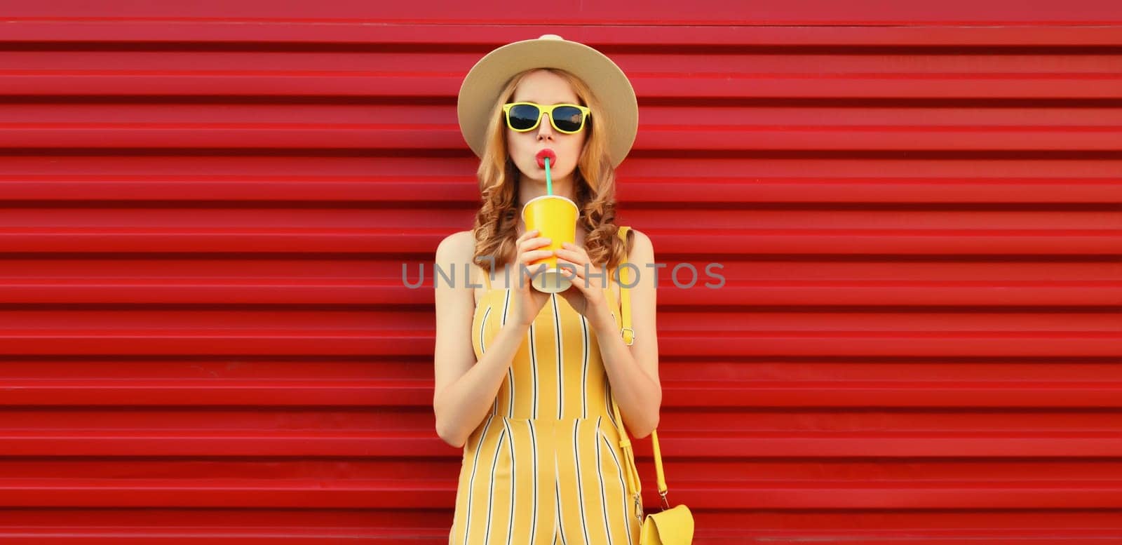 Beautiful stylish happy young woman drinking fresh juice holding cup wearing summer hat, yellow outfit posing on red wall background