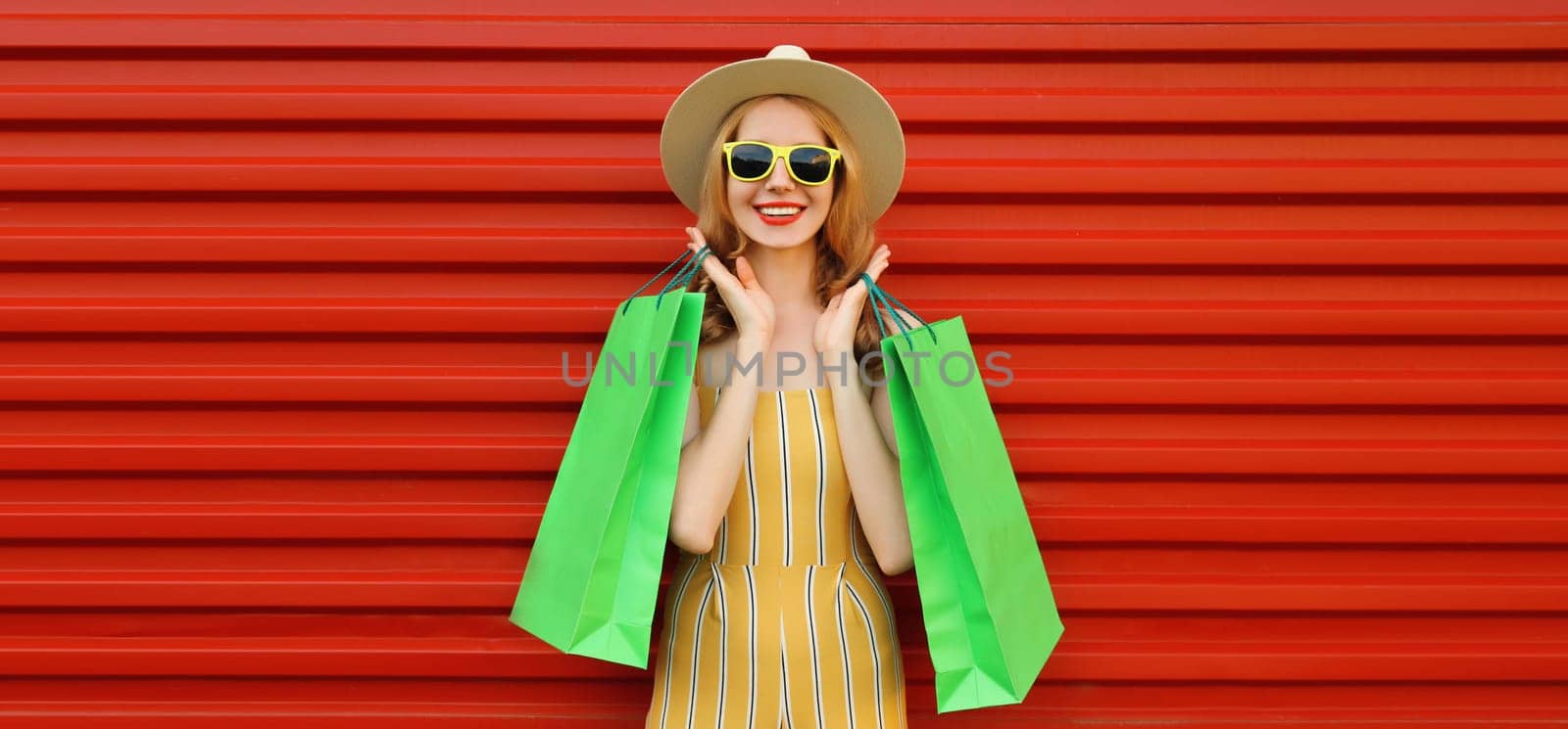 Stylish beautiful happy smiling young woman posing with bright green shopping bags wearing summer straw hat on red wall background