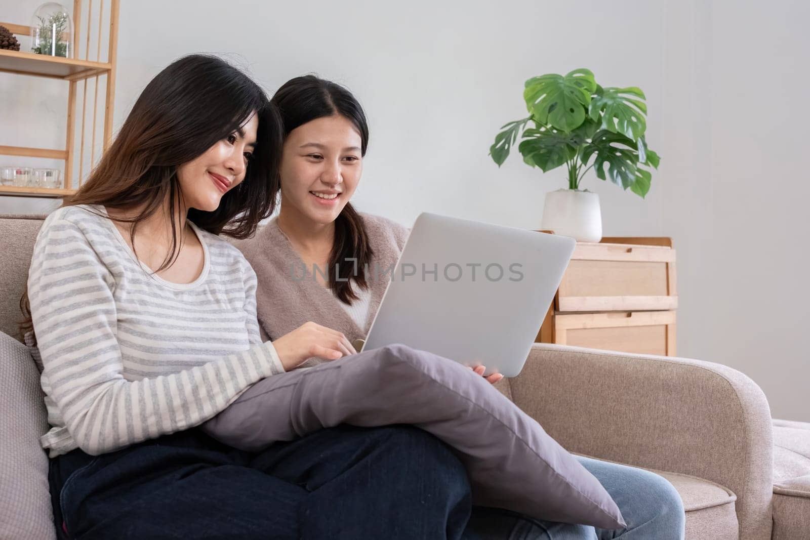 Asian lesbian couple enjoying leisure time at home on the couch with a laptop. Concept of love, companionship, and modern lifestyle in a cozy indoor setting.