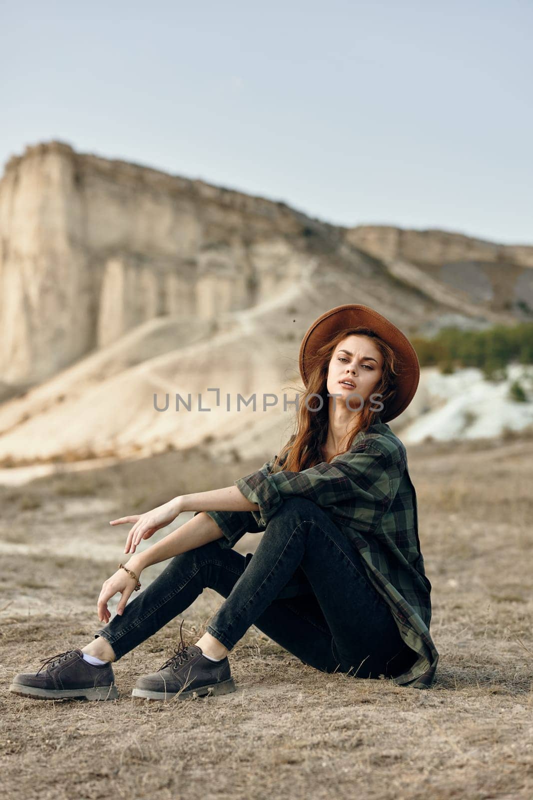 Serene young woman in hat sitting peacefully in front of majestic mountain landscape on sunny day