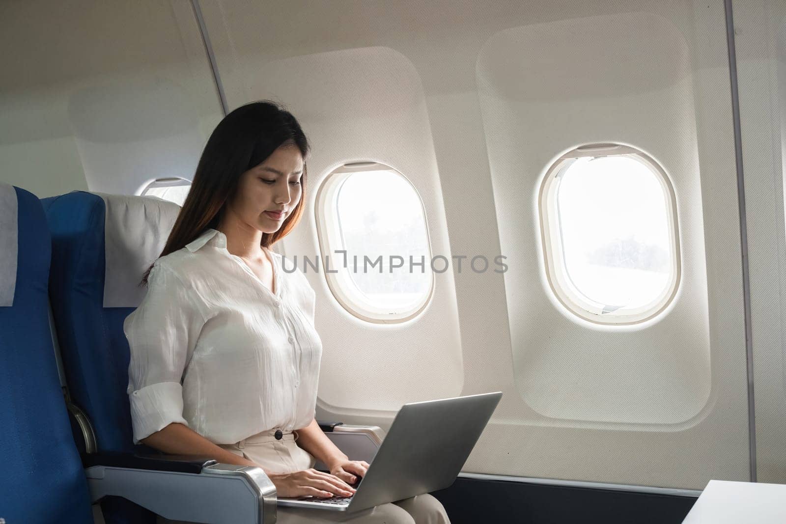 Asian woman working on laptop in airplane seat. Concept of air travel, remote work, and in-flight productivity.