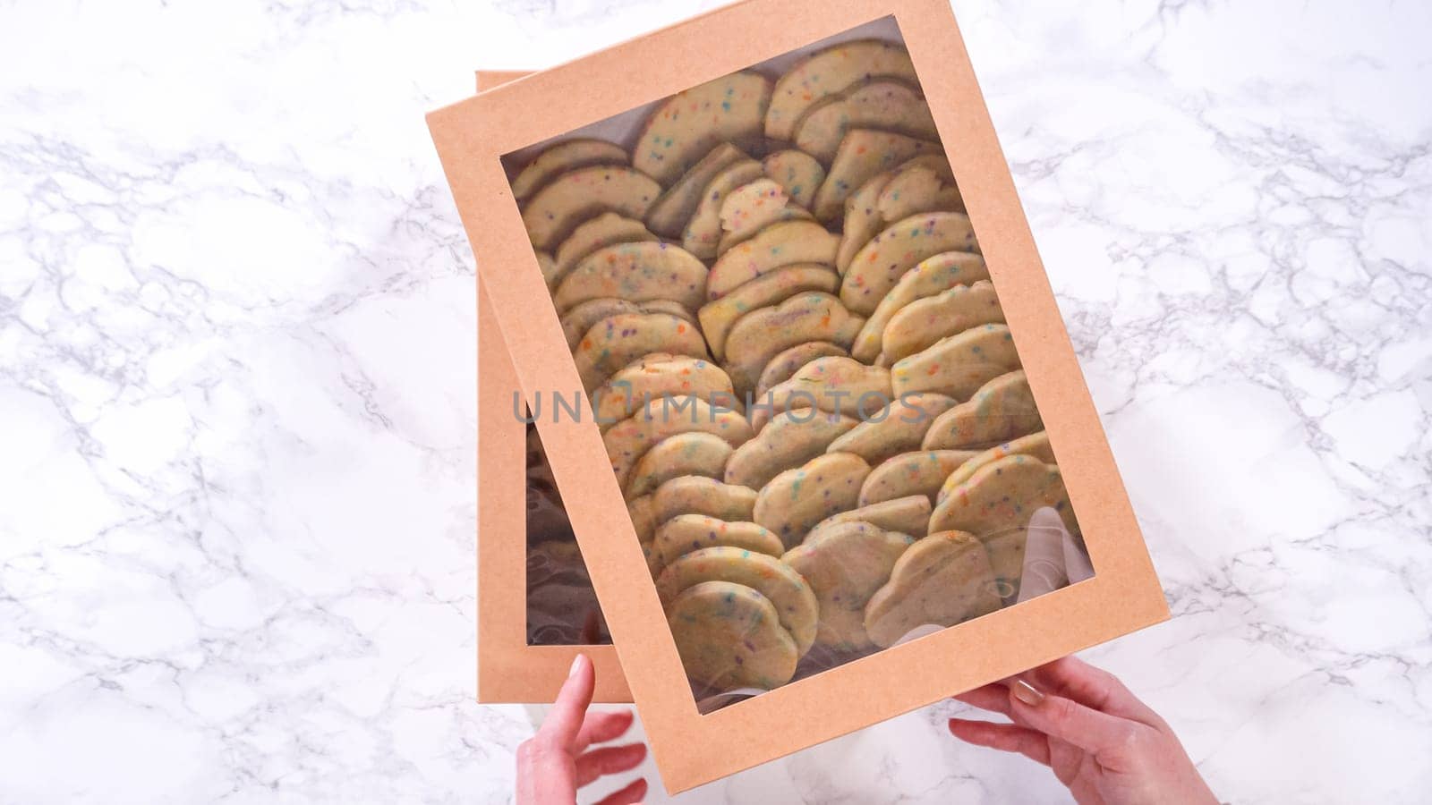 Flat lay. With precision, the woman is carefully arranging the sugar cookies, filled with dough-mixed sprinkles, into a rustic brown paper box.