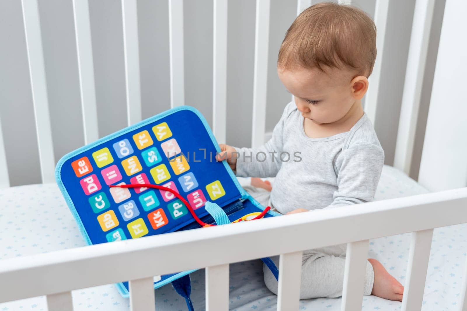 One year old baby playing with montessori busy book sitting in crib