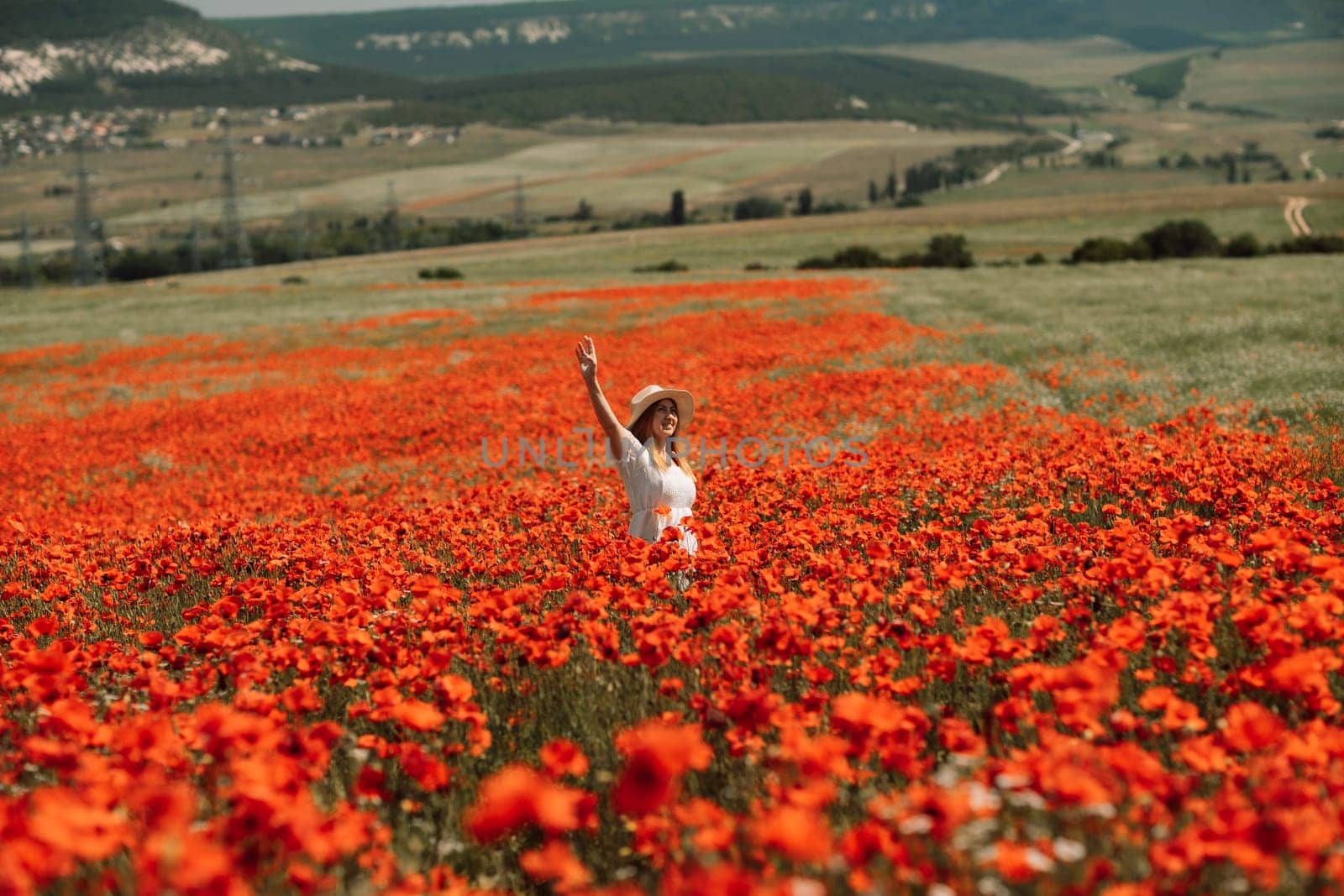 Field poppies woman. Happy woman in a white dress and hat stand through a blooming field of poppy raised her hands up. Field of blooming poppies