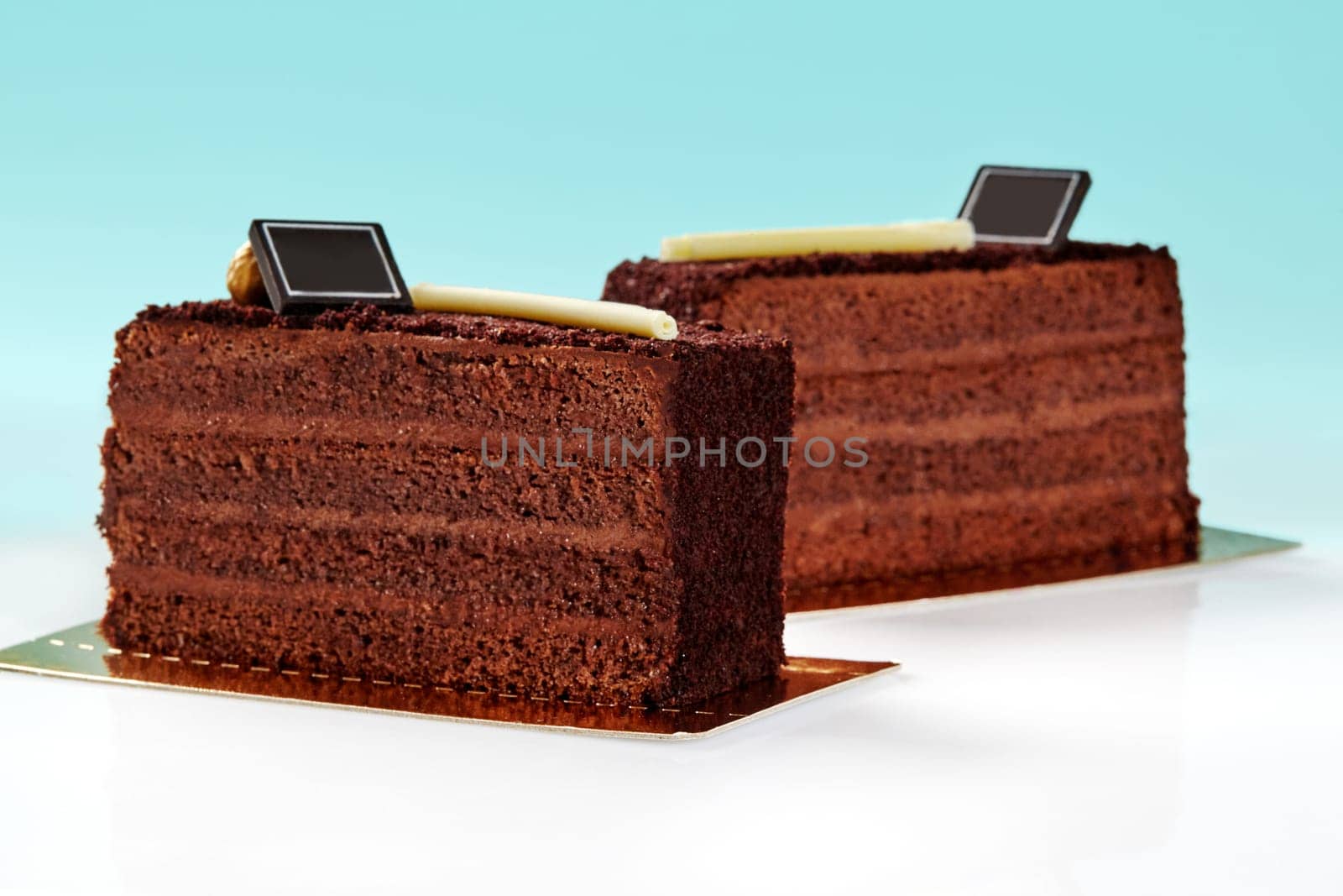 Two enticing slices of rich chocolate cake topped with crunchy streusel decorated with white chocolate stick, branding plaque and caramelized hazelnut on pastel blue background