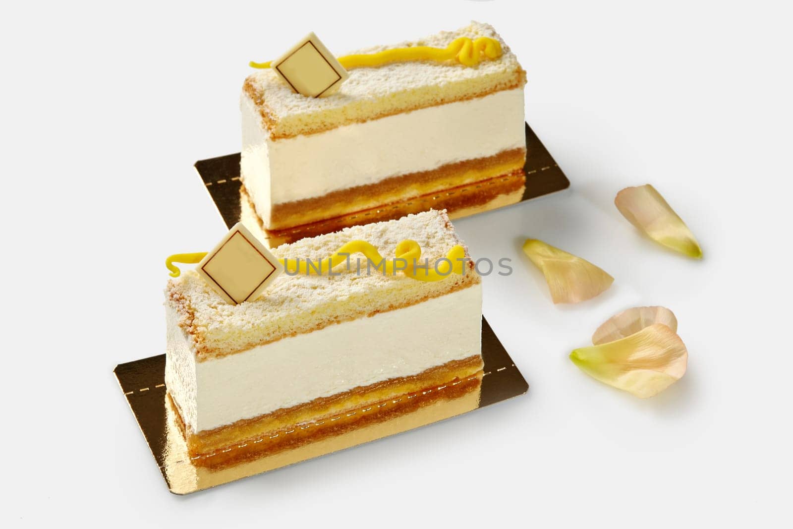 Slices of tempting birds milk sponge cake filled with airy creamy souffle decorated with signature chocolate plaque and yellow mastic swirl, on white background with flower petals